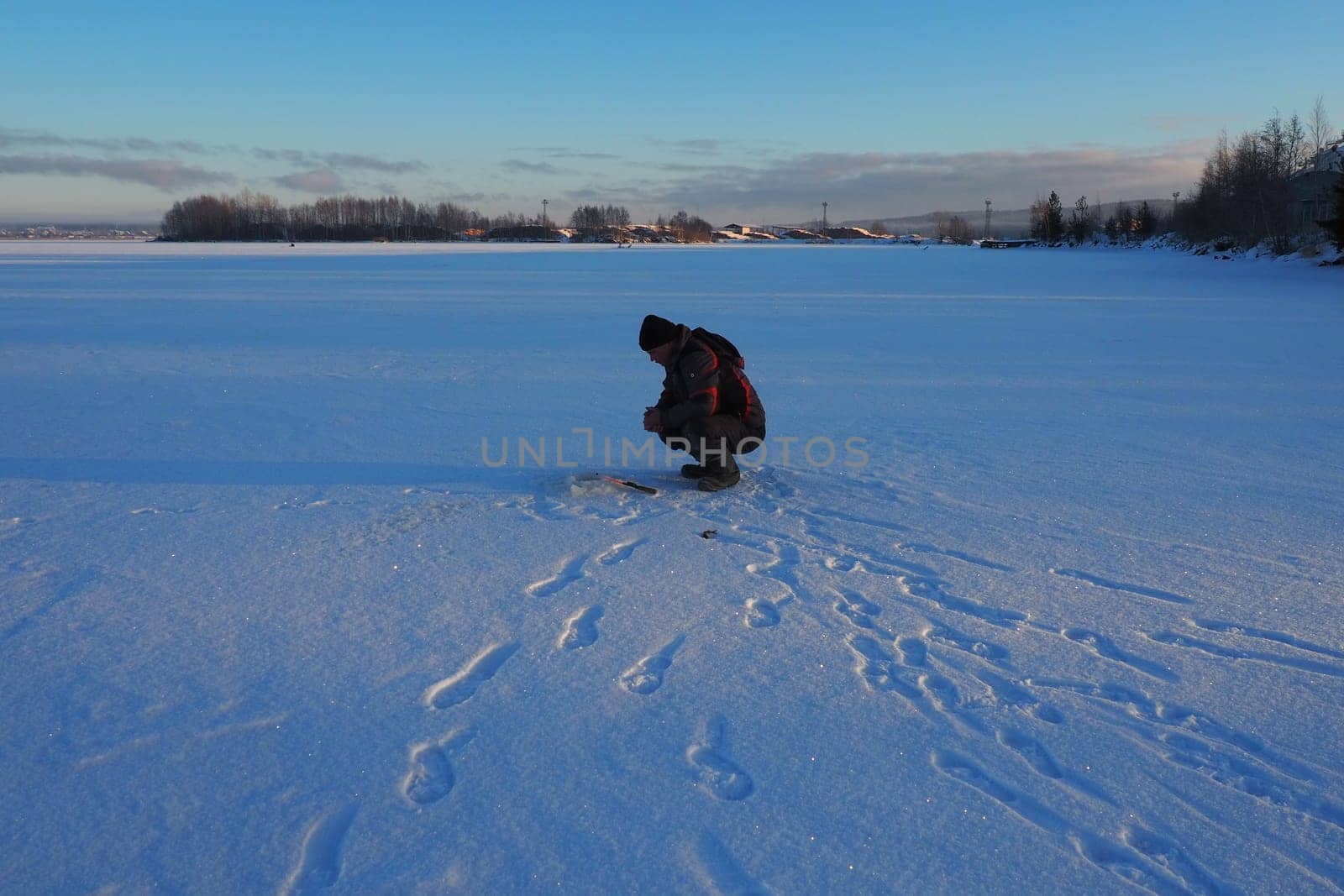 Fisherman on winter fishing. A man sits near a hole in a frozen lake waiting for a bite. Snow on ice. Bottom fishing for perch. Frosty January evening