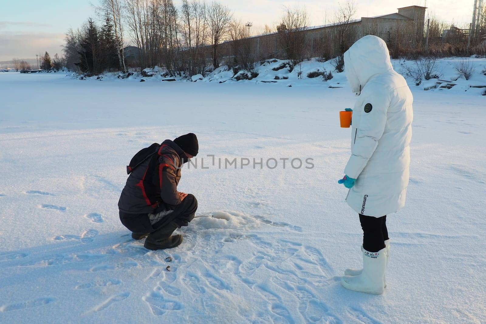 Logmozero, Karelia, Russia, January 08, 2022. Fisherman and fisherwoman on winter fishing. A man sits near a hole in a frozen lake and waits for a bite. Snow on ice. Bottom fishing for perch.