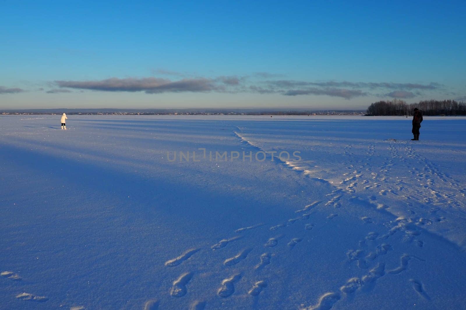 Fisherman and fisherwoman on winter fishing. A man sits near a hole in a frozen lake and waits for a bite. Snow on ice. Bottom fishing for perch. Frosty January evening. Catching perch on a bloodworm