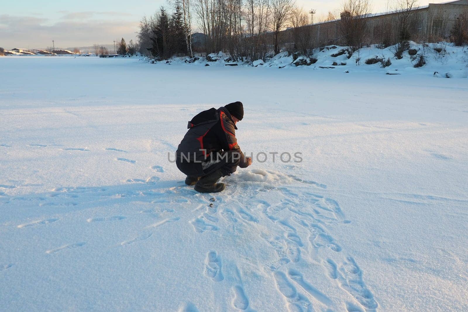 Fisherman on winter fishing. A man sits near a hole in a frozen lake waiting for a bite. Snow on ice. Bottom fishing for perch. Frosty January evening