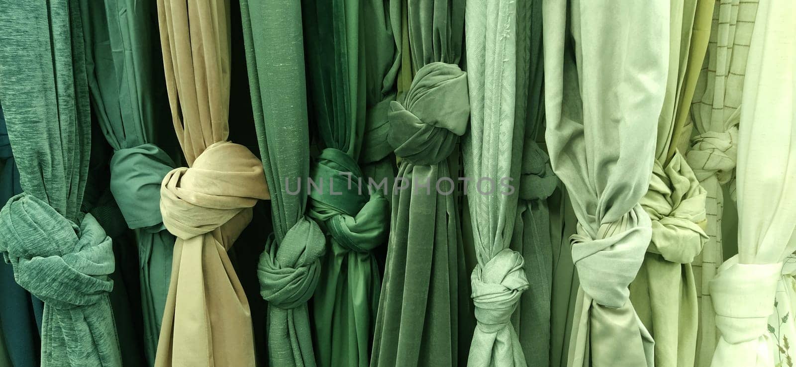 Curtain and fabric for drapery and interior decoration. Samples of fabrics in the store are hung vertically and tied in knots. Green, beige colored and white curtains in the showcase. Store shelves