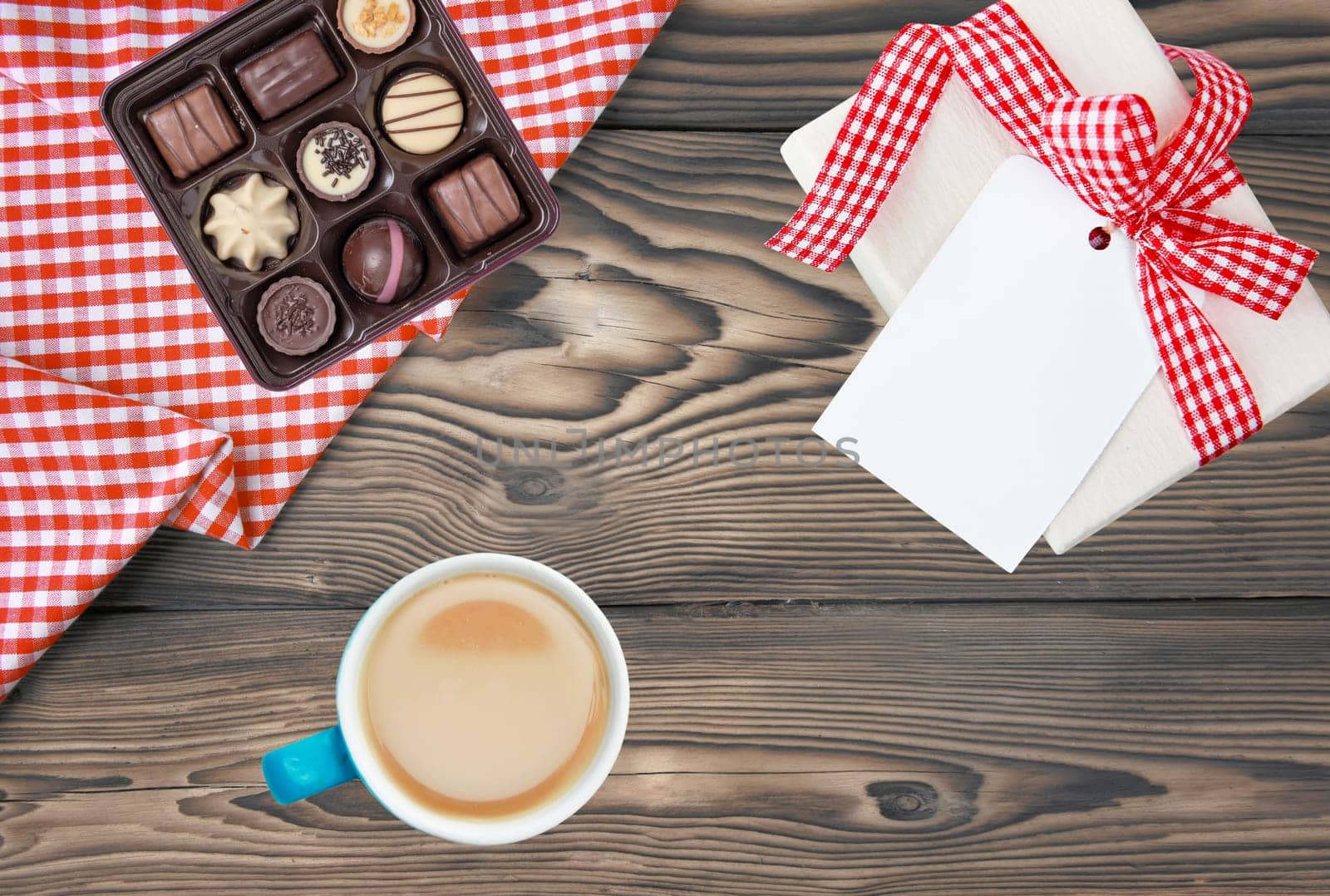 Coffee mug, present box with blank paper tag and box of fine chocolate candies on rustic wooden table. Top view. Breakfast on Valentine, Mother or Womens Day. Copy space. Card concept.