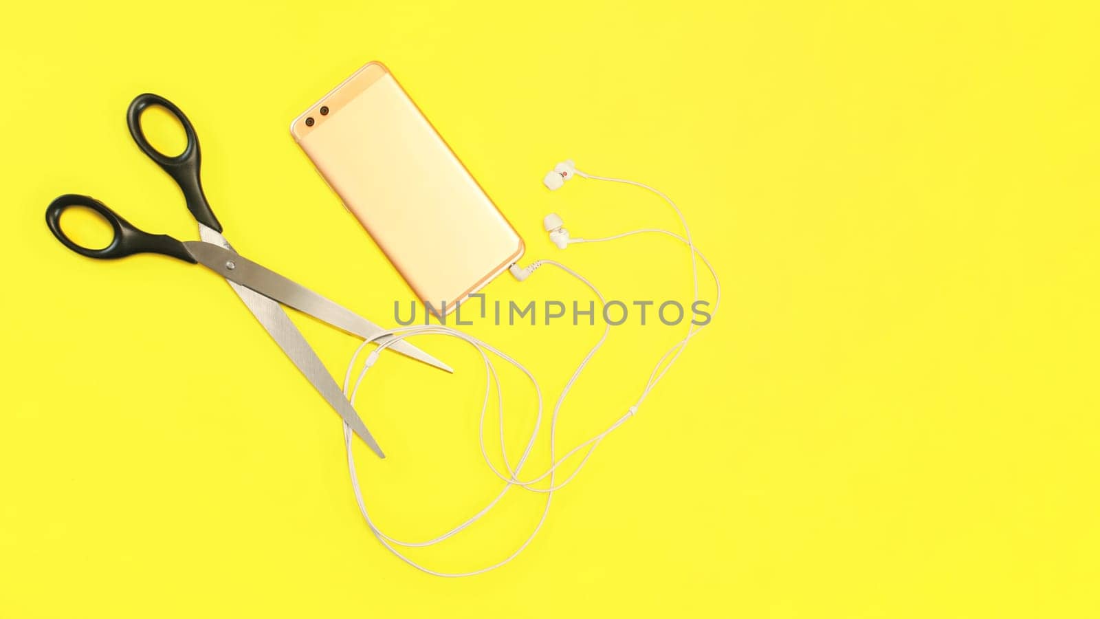 Top view on rose gold cell phone with ear buds cable to be cut with large scissors on yellow background.