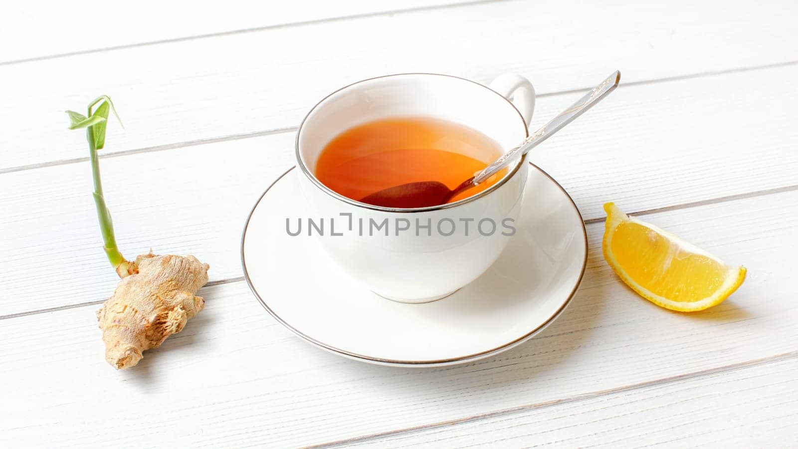 White porcelain cup with hot amber tea, silver spoon in it, dry ginger root green sprout and piece of lemon at side.
