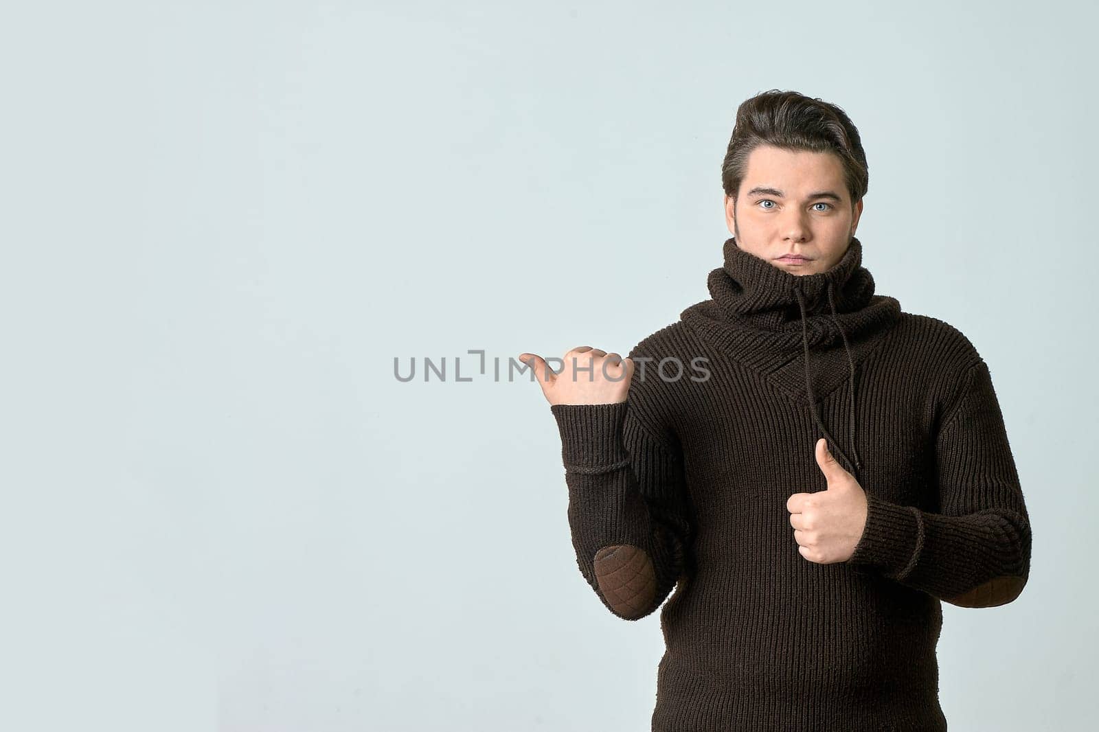 a man in a brown sweater and a bright color points to something and perfectly, showing an empty space with his thumb, shows everything on a light background, copy space. High quality photo