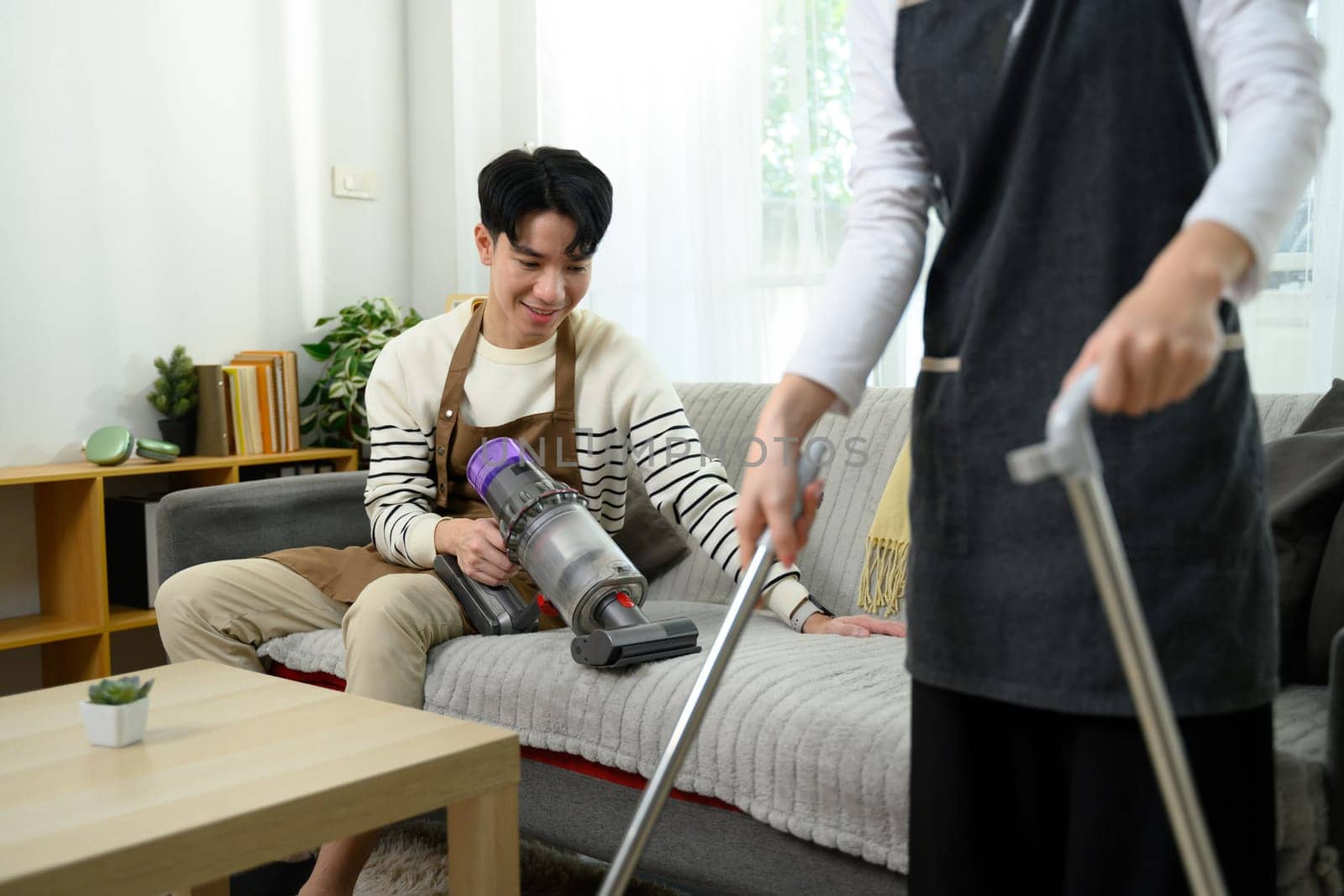 Cheerful young couple wearing aprons work together to clean their living room.