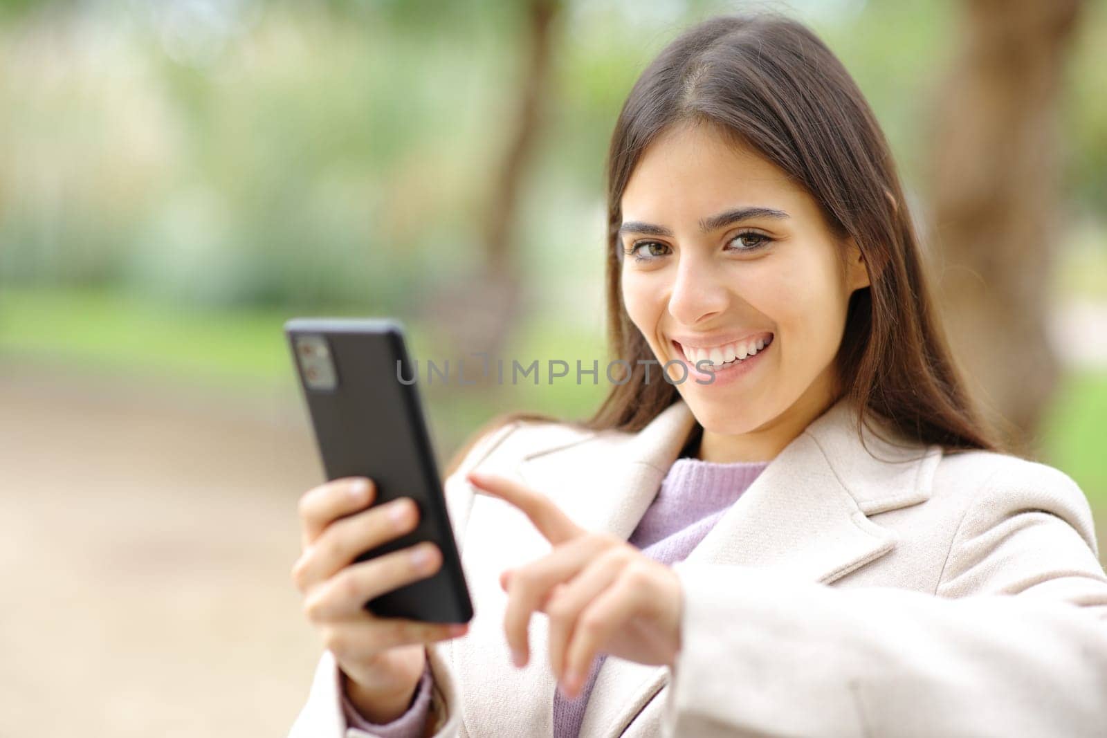 Woman in winter looks at you holding phone in a park