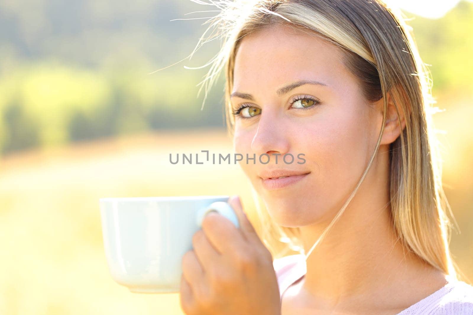Satisfied woman looks at you holding coffee cup in a field