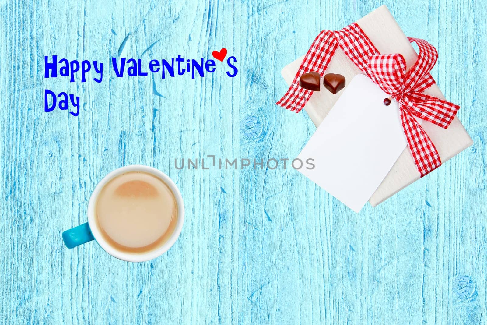 Coffee mug, present box with chocolate hearts and blank paper tag on blue wooden desk with Valentine text. Breakfast on Valentine day. Happy Valentines Day greeting card.