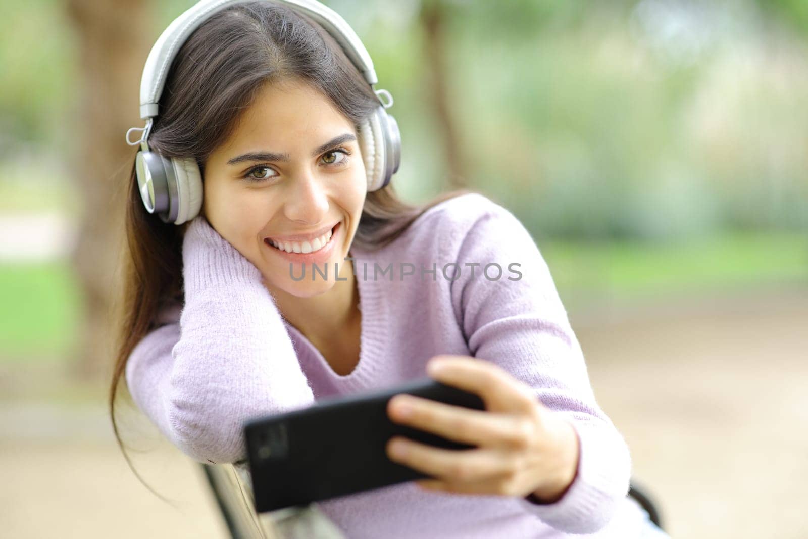 Happy woman listening to music looking at you in a park