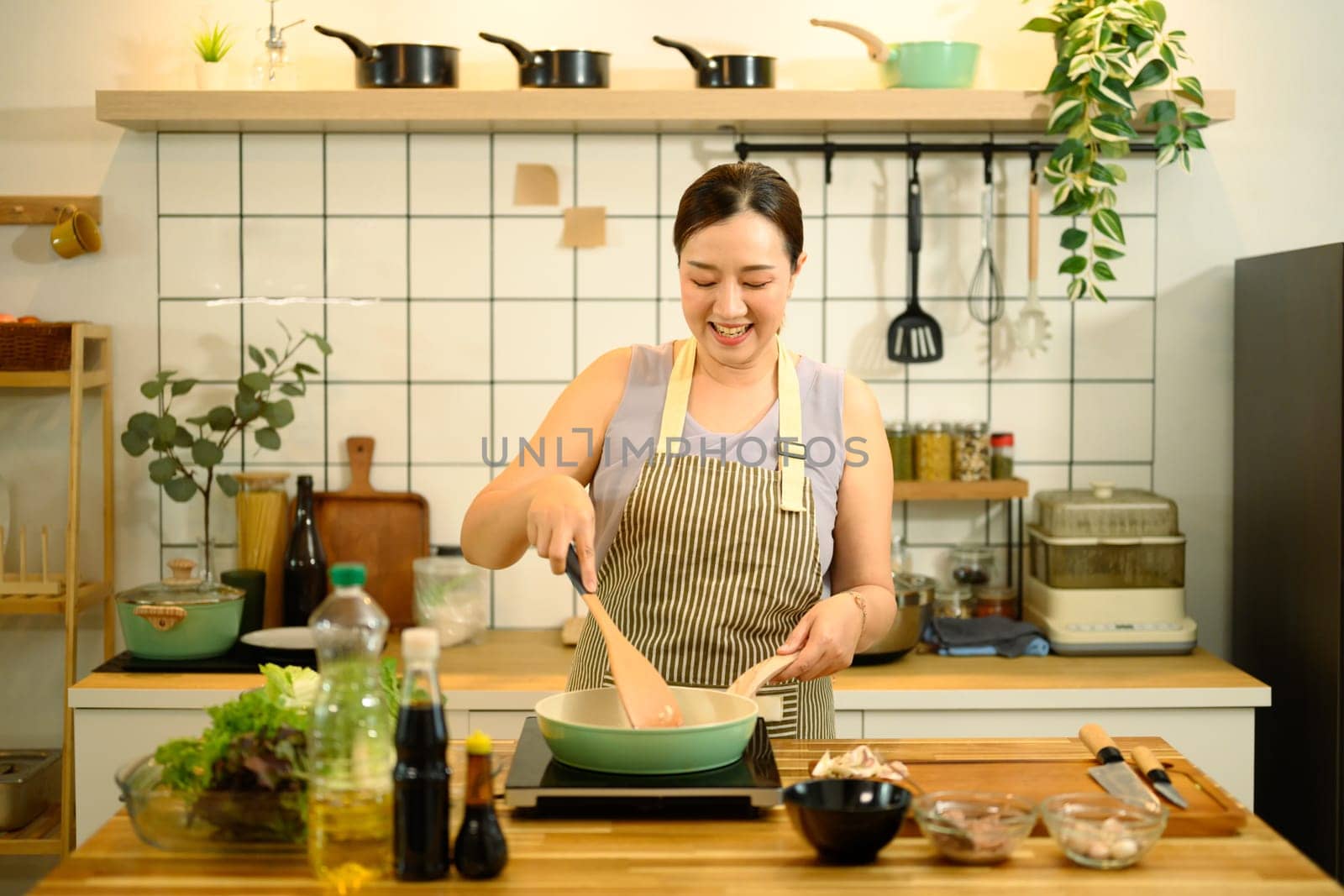 Smiling middle aged woman stirring ingredients in frying pan while preparing meal in cozy kitchen.