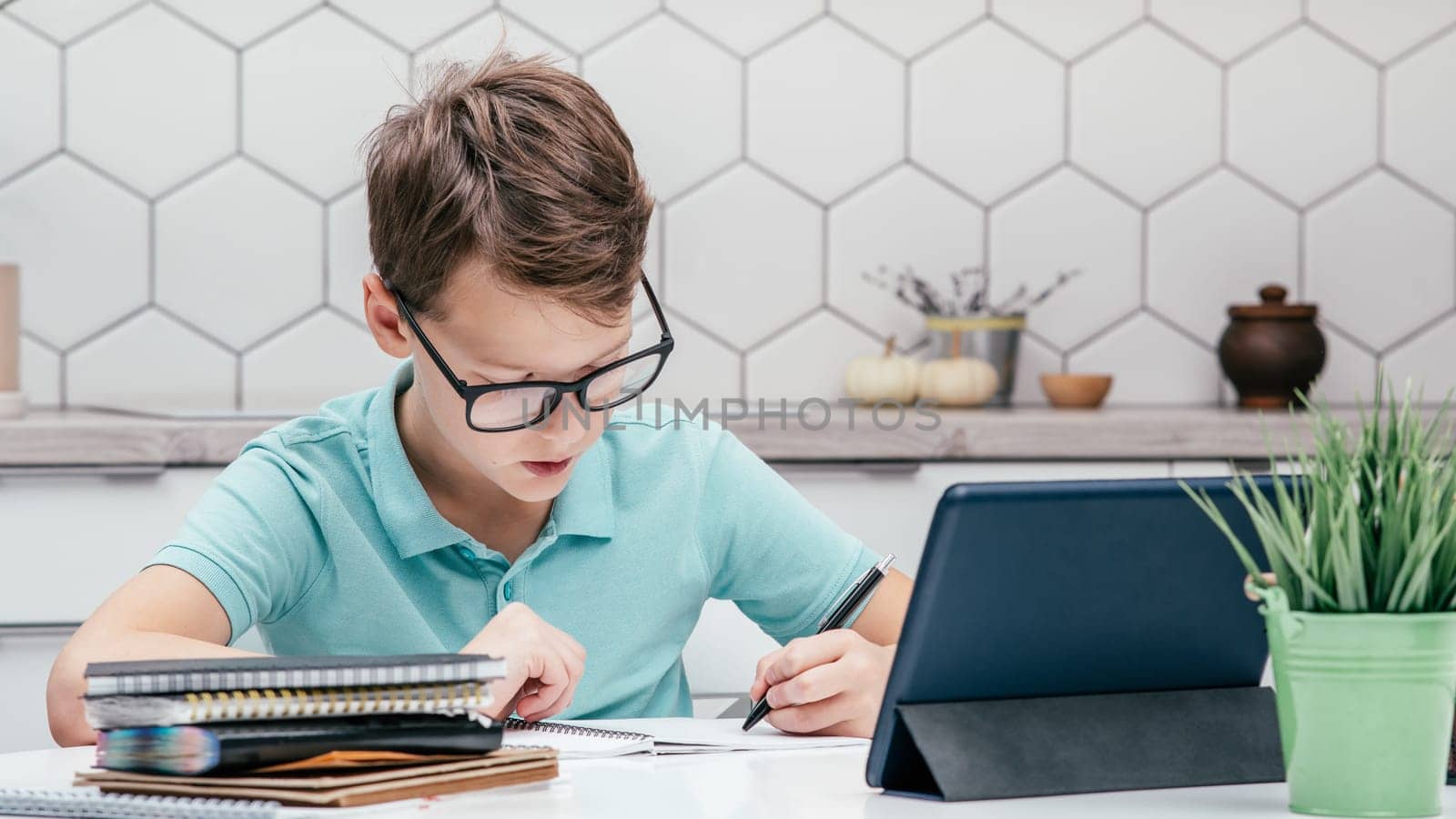 Portrait of preteen concentrated boy pupil wearing blue T-shirt, glasses, sitting at desk near notebooks, tablet, doing homework with left hand, writing on notebook. Distant learning, education.