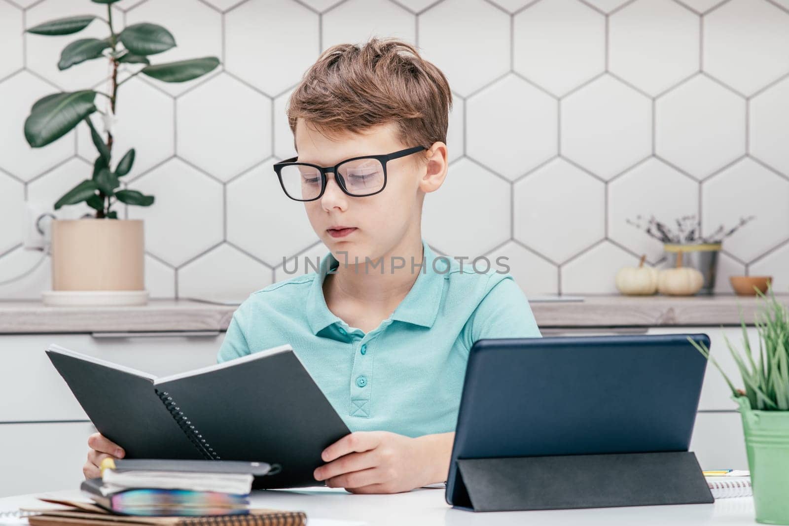 Portrait of young preteen serious boy wearing blue T-shirt, glasses, sitting at desk near notepads, looking at book near tablet, houseplant, having online lesson, reading. E-learning, technology.