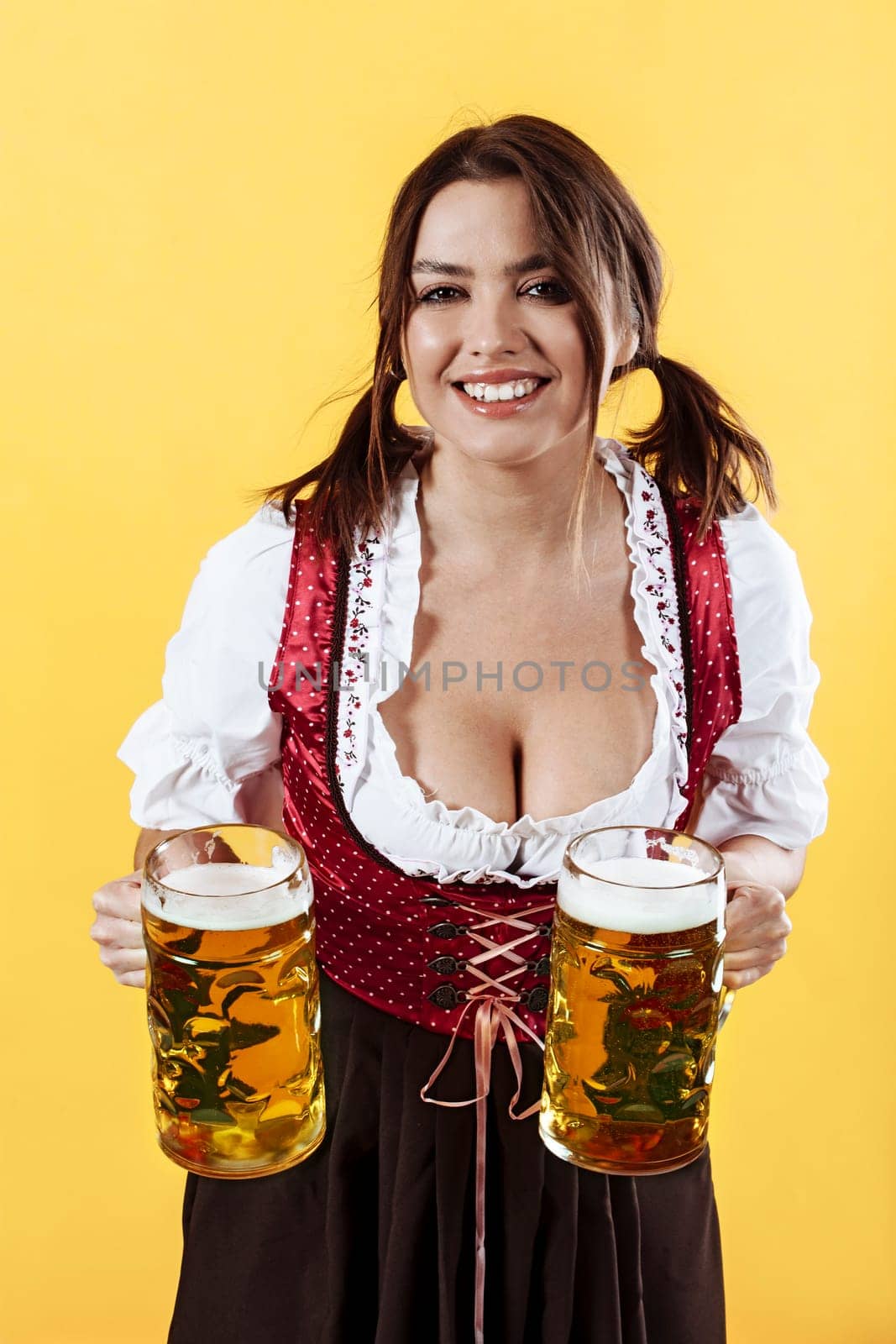 Beautiful Oktoberfest server wearing traditional dirndl and holding two mass beer steins over yellow background.