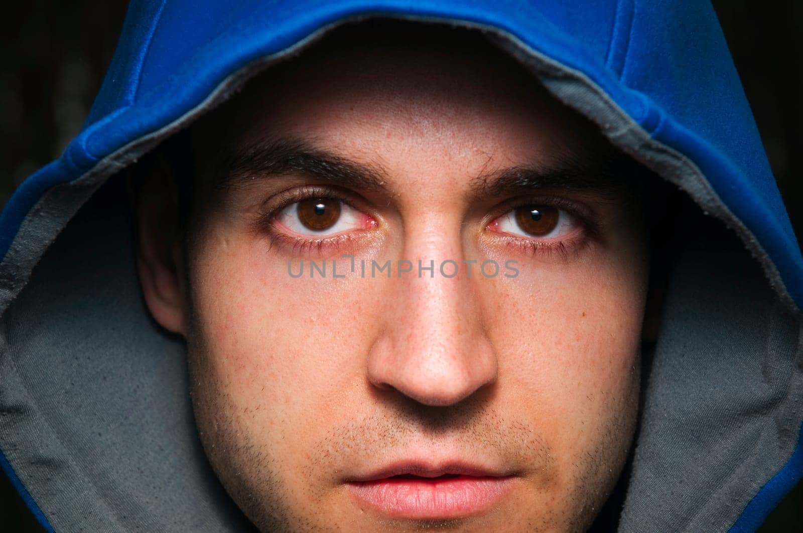 Frontal face portrait of a blue hooded young man looking directly and piercingly at the camera.