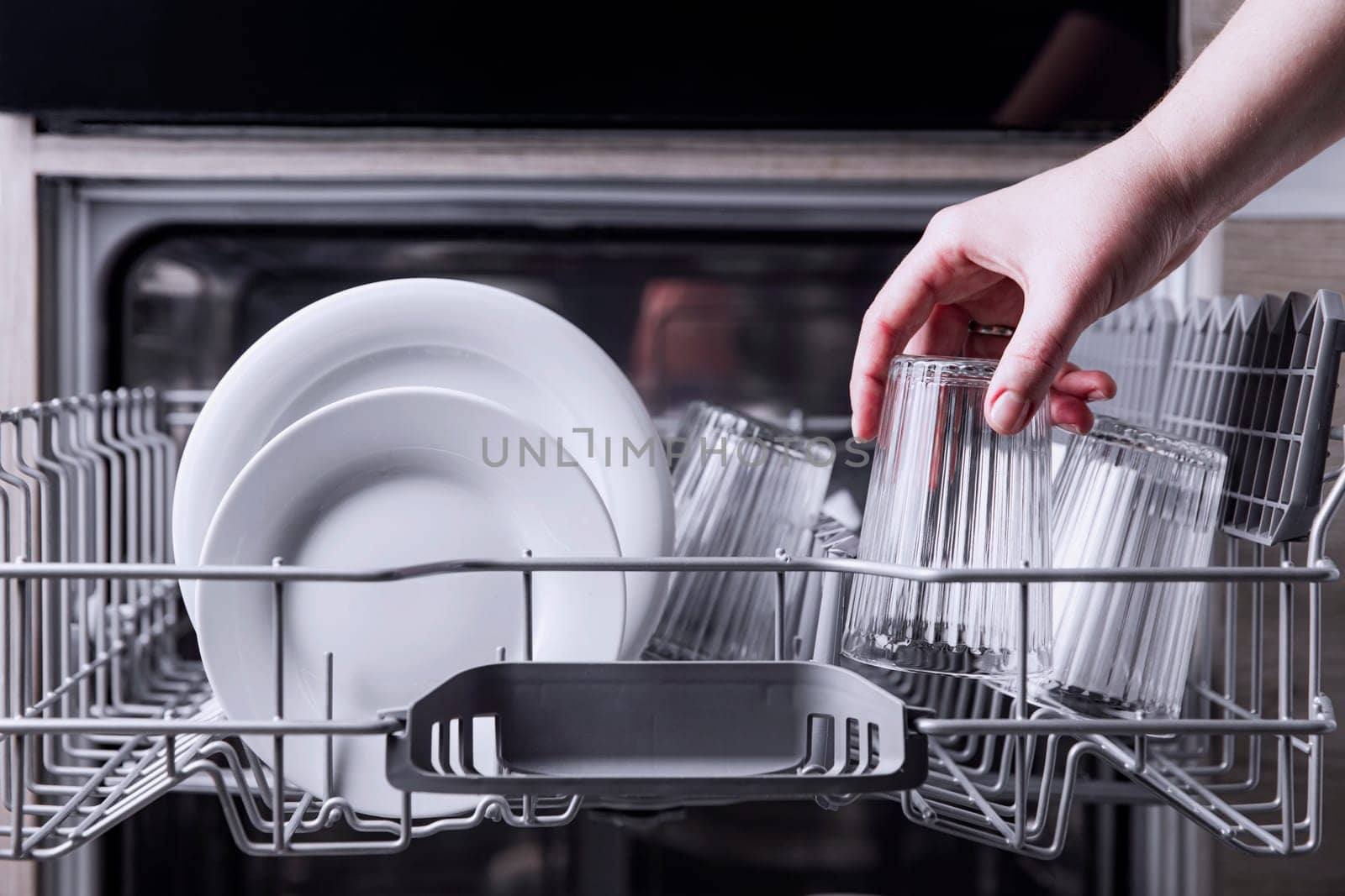 Female hand loading dished, empty out or unloading dishwasher with utensils. Kitchen appliances, lifestyle view. Woman puts a plate in the dishwasher or takes from it. Housewife does her housework