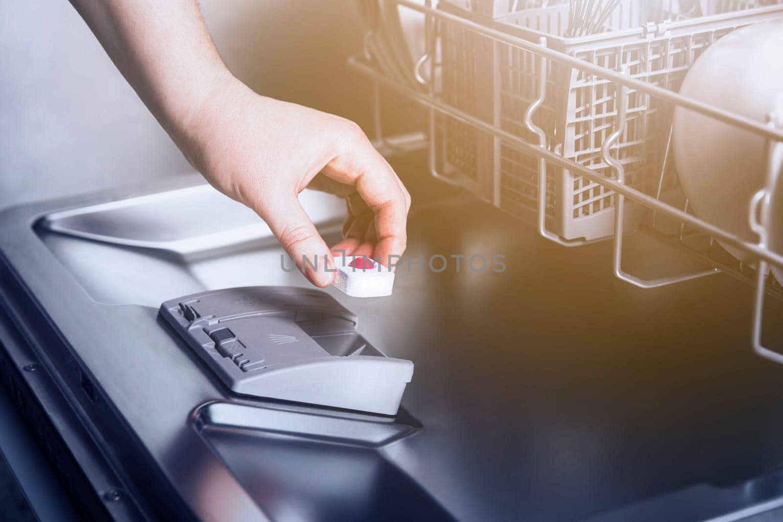 Close-up of female hand inserting dishwasher tablet into open automatic stainless built-in dishwasher machine with dirty utensils inside in modern home kitchen. Household, housekeeping domestic life