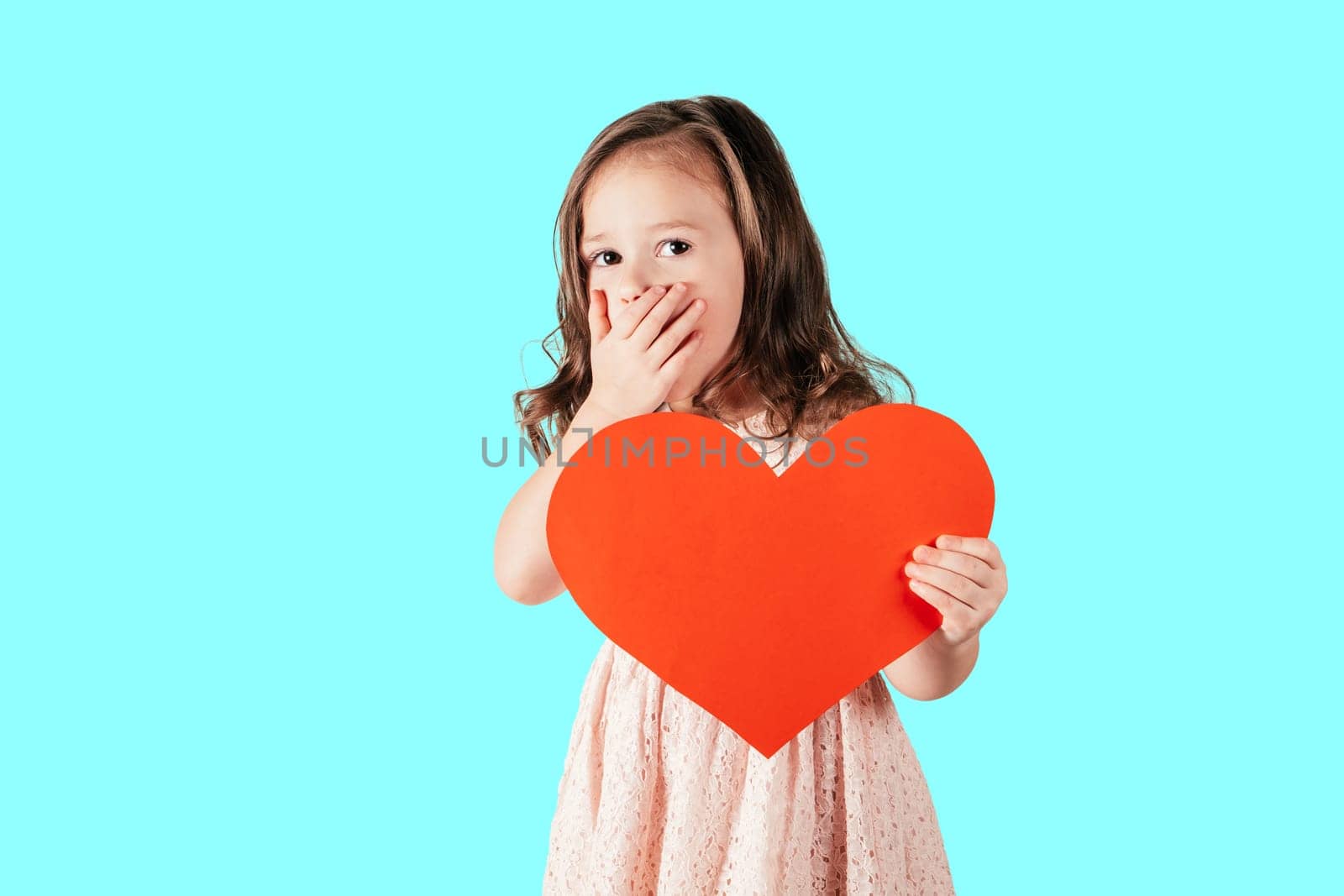Portrait of little girl with long curly hair hold in hand red paper heart putting hand over mouth, isolated on blue background. Valentines day, birthday, Mother's day concept. Copy space