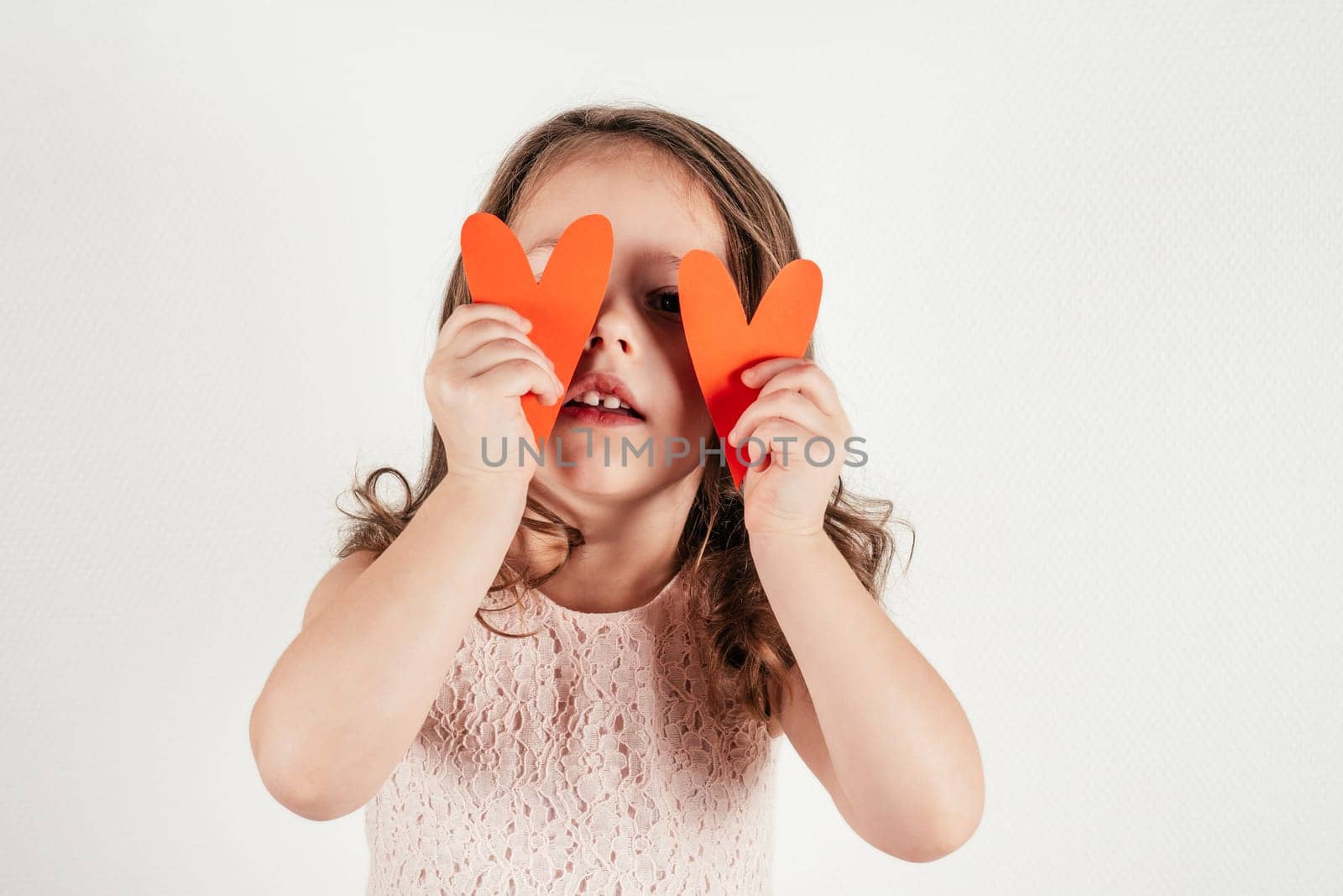 Portrait of beautiful little girl with long curly hair hold in hands two red paper hearts covering eyes on white background. Family, love, holidays, birthday, Mother's day concept. Copy space. Mock up