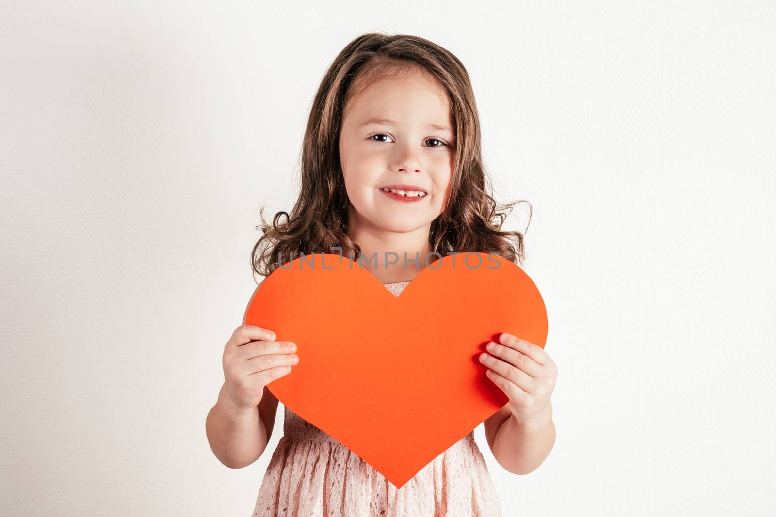 Portrait of little girl with long curly hair hold in hands red paper heart smiling on white background. Congratulations on family holidays, Valentines day, birthday, Mother's day concept. Copy space.