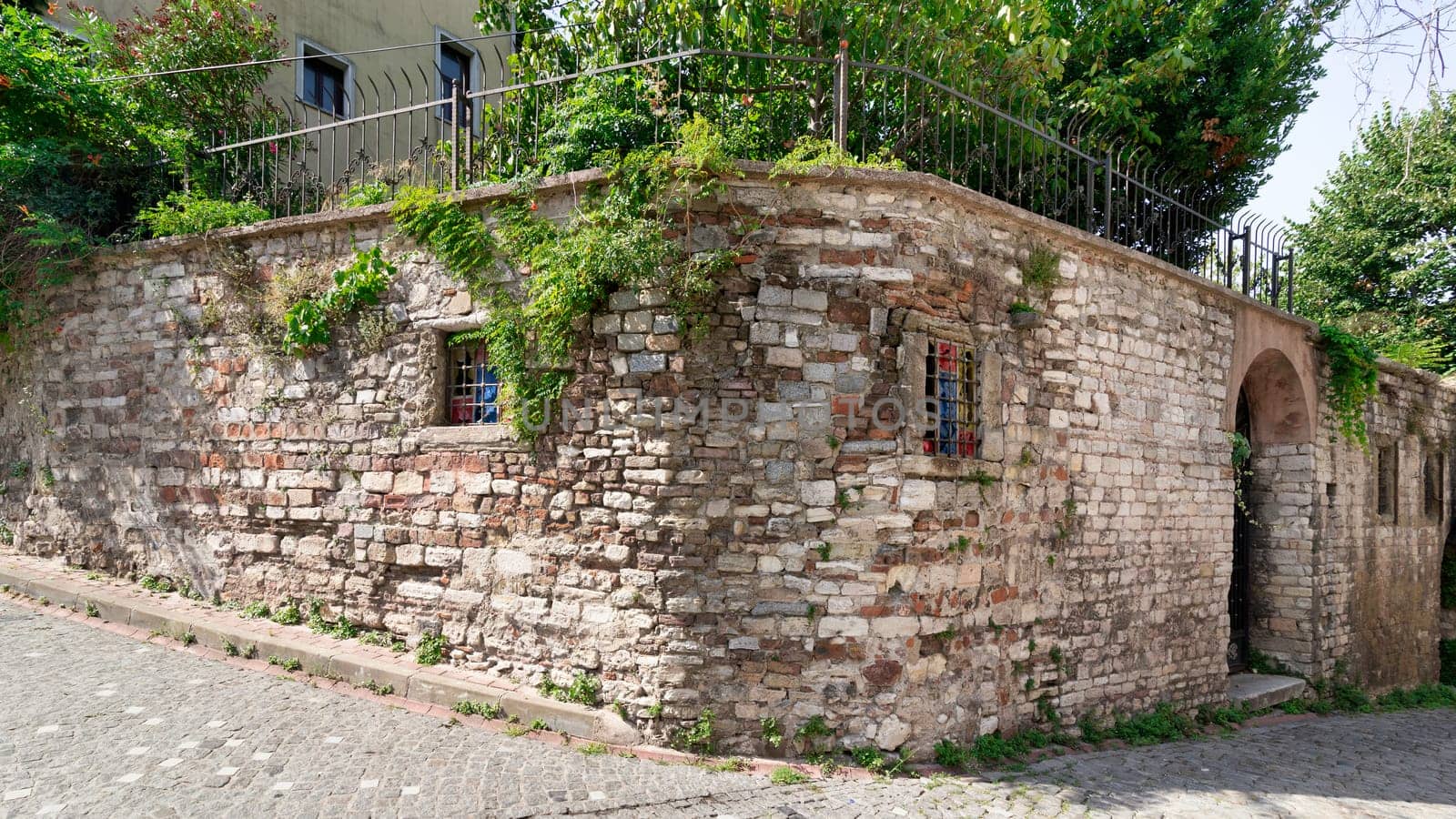 Old abandoned building, with weathered stone wall, and wrought iron windows, partially covered with climber plants, in a narrow alley with cobblestone floor, Balat district, Istanbul, Turkey