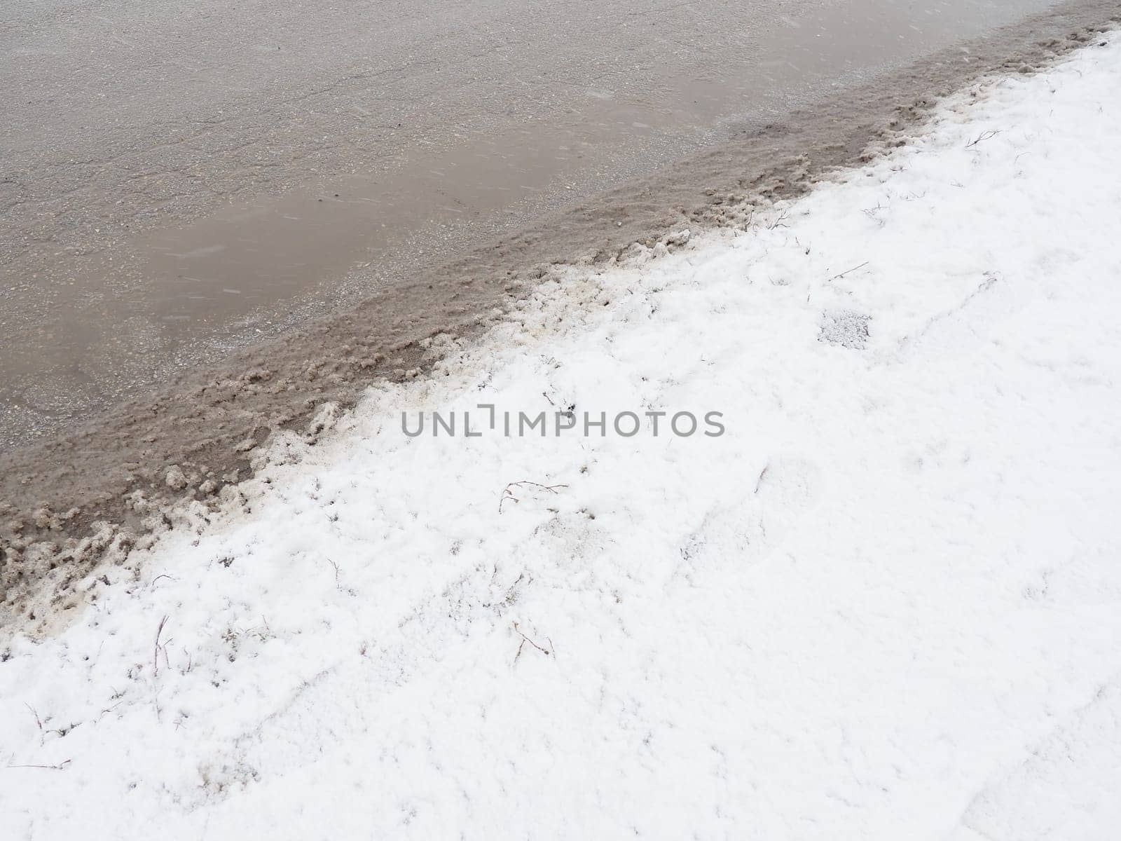 Roadside with a snowdrift. Difficult driving conditions. Wet slippery pavement with footprints. Dangerous ice. Muddy weather in winter. Car tire tracks. The work of municipal services for cleaning.