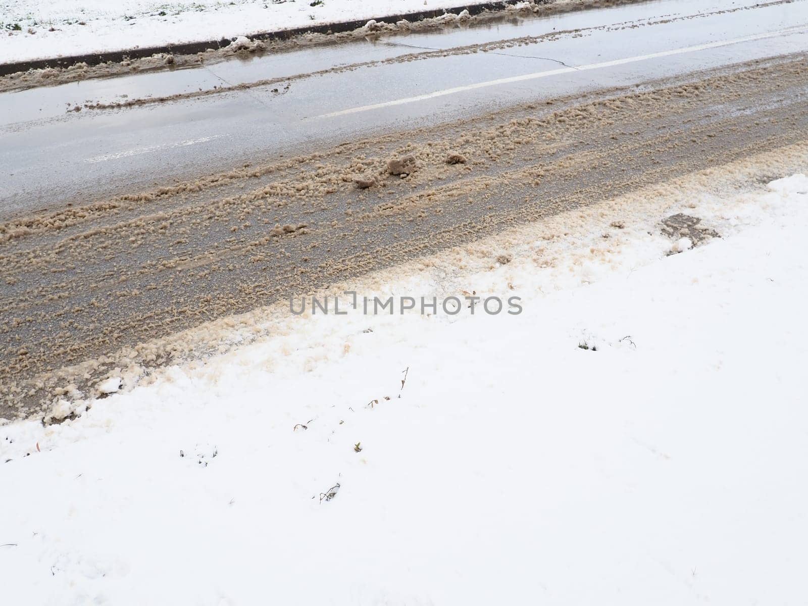 Roadside with a snowdrift. Difficult driving conditions. Wet slippery pavement with footprints. Dangerous ice. Muddy weather in winter. Car tire tracks. The work of municipal services for cleaning.