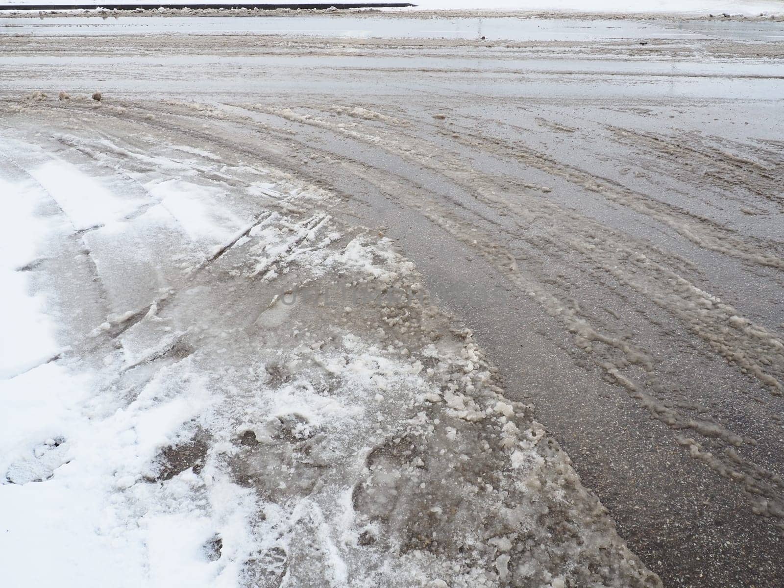 Roadside with a snowdrift. Difficult driving conditions. Wet slippery pavement with footprints. Dangerous ice. Muddy weather in winter. Car tire tracks. The work of municipal services for cleaning.