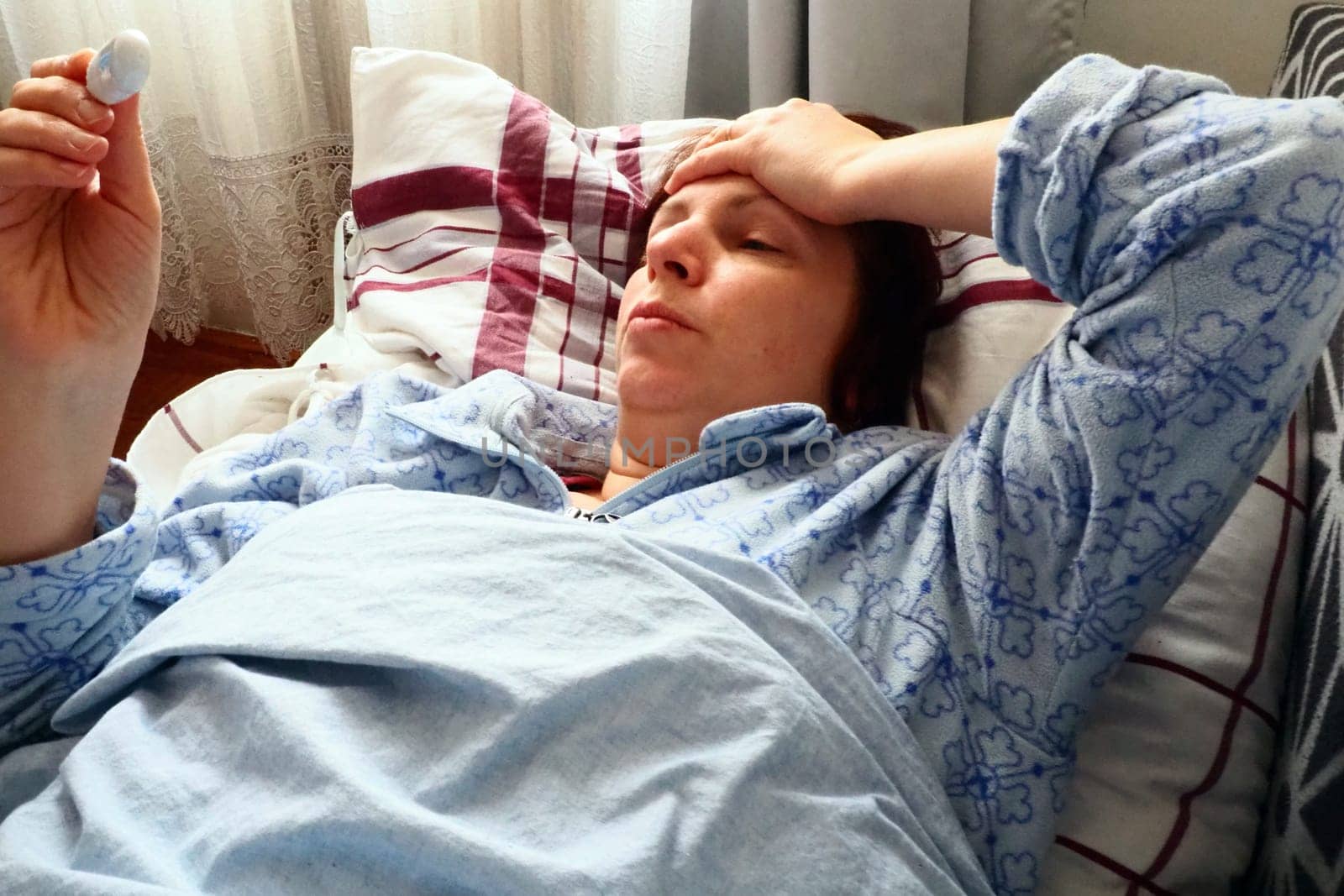 Sick woman in bed. Headache, runny nose and cold. Paleness of the face and faintness of the body. Feeling unwell. The patient lies on a pillow, holds a thermometer and measures the temperature