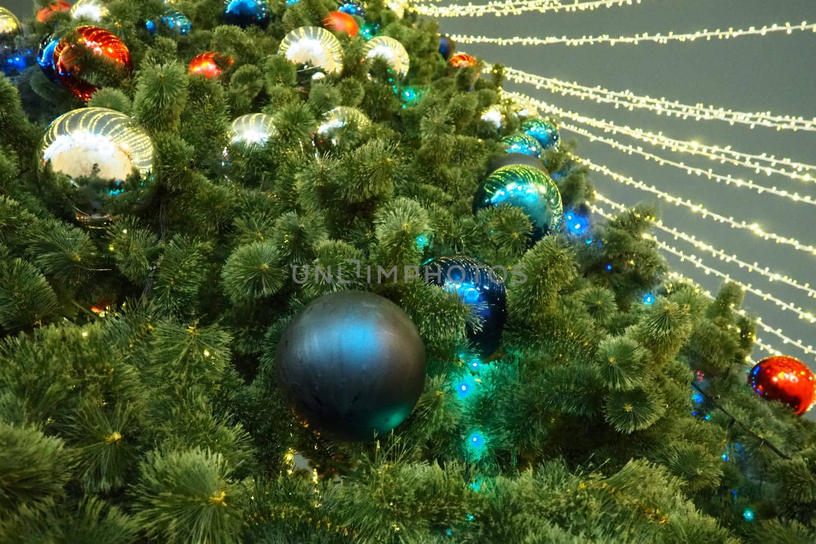 Multi-colored glass shiny balls on the New Year's city tree. Glowing garlands hang over the Christmas tree. Outdoor winter decoration. Festive design of the city square. Red, blue and yellow ball.