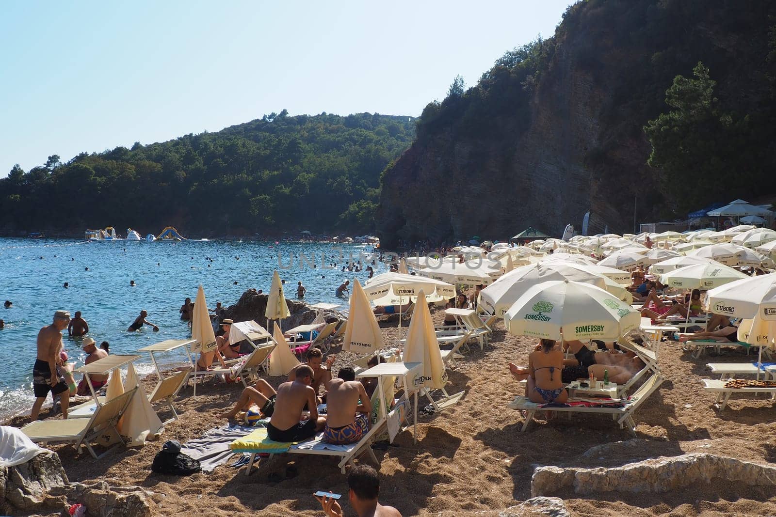 Budva, Montenegro, Mogren beach. August 15 2022 Sand and pebble beach near high cliffs. Beach umbrellas, cafes, deck chairs. People relax, swim, enjoy and sunbathe. Tourism business in the Balkans