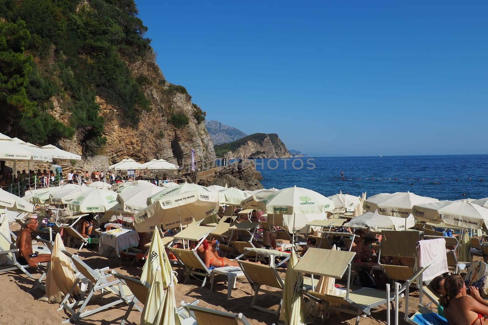Budva, Montenegro, Mogren beach. August 15 2022 Sand and pebble beach near high cliffs. Beach umbrellas, cafes, deck chairs. People relax, swim, enjoy and sunbathe. Tourism business in the Balkans