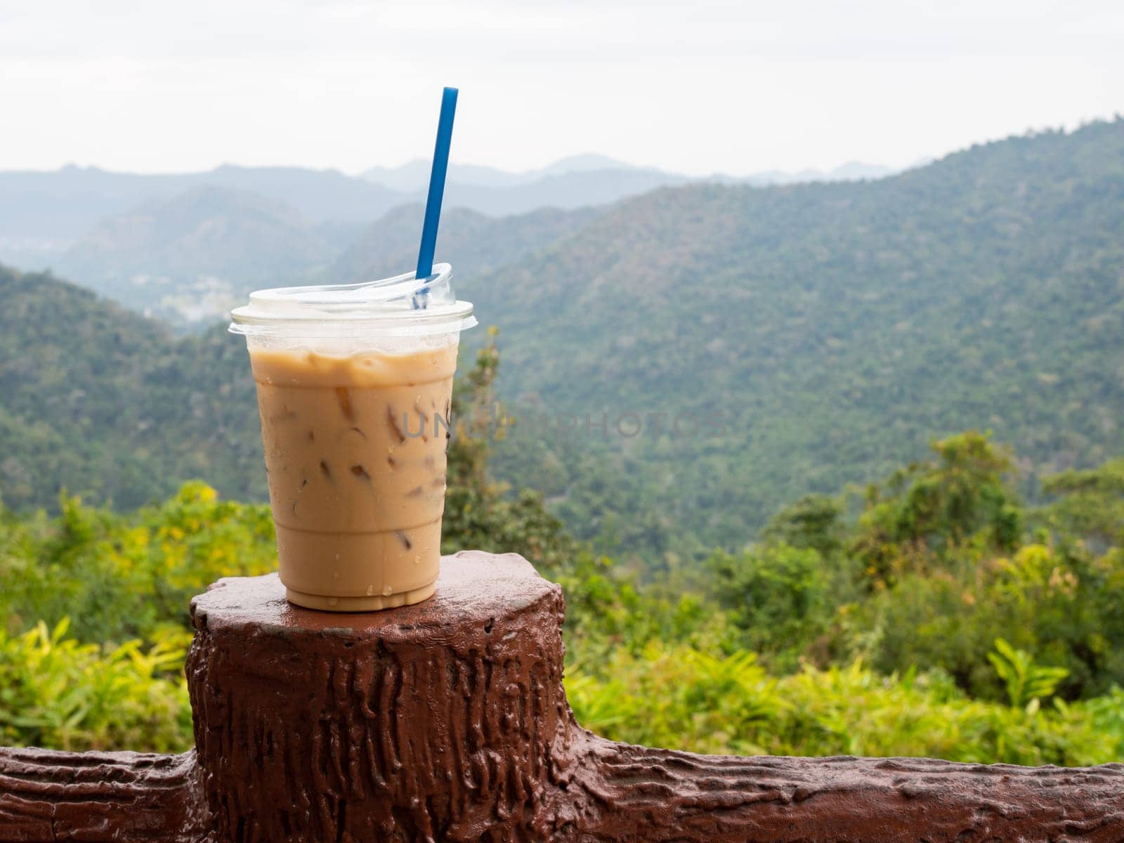 A glass of iced coffee is placed on the fence against a backdrop of mountains and sky.