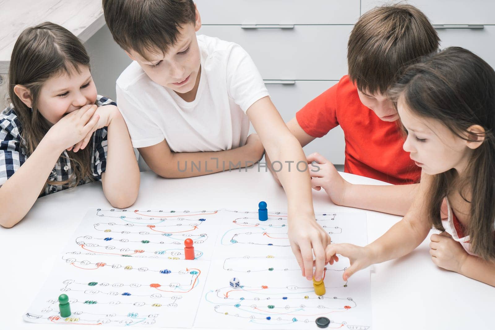 Top view of group of children friends classmates sitting at white table, playing board game tabletop game, moving colorful figures on white board with numbers, tracks. Board games, entertainment.