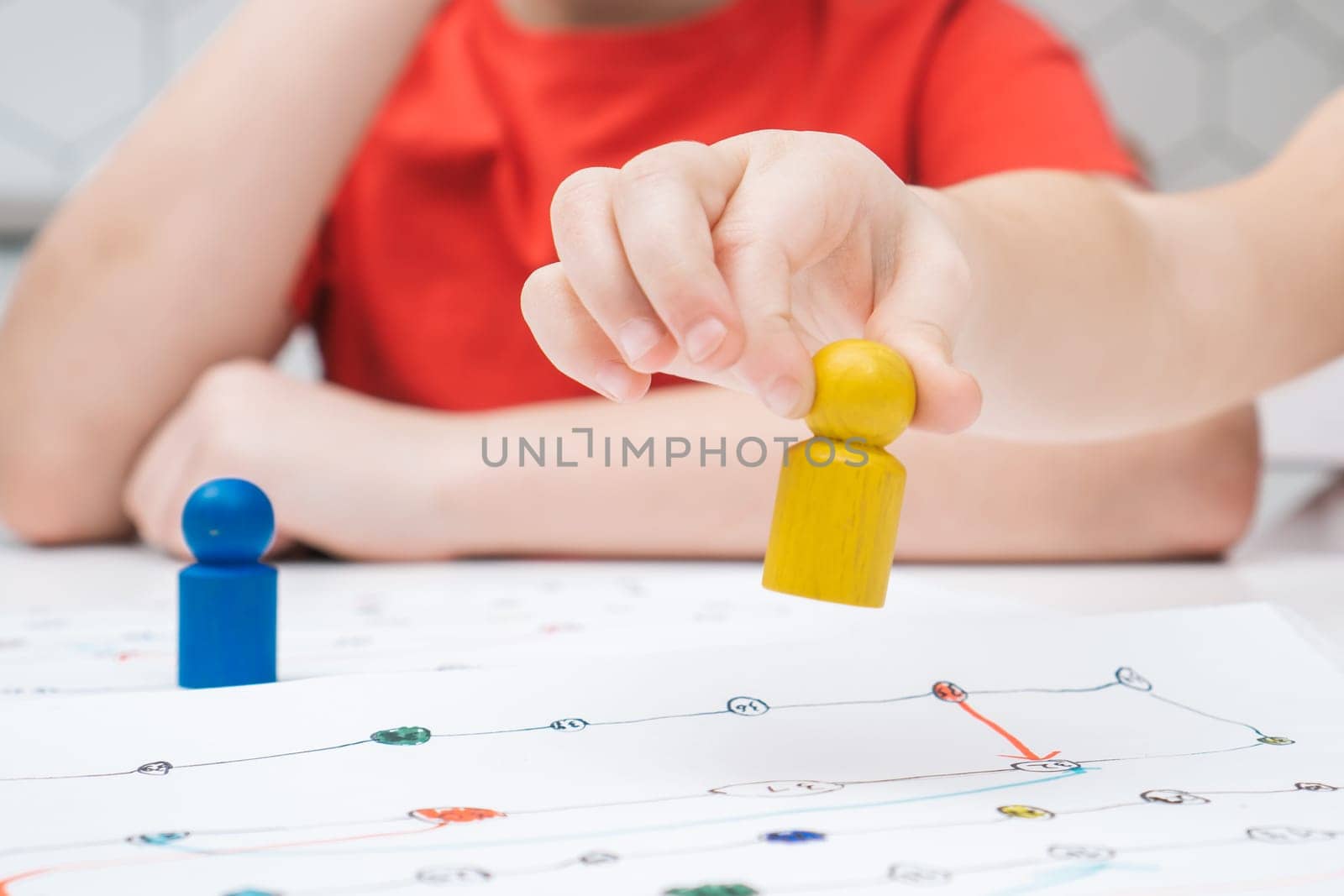 Cropped photo of group of children sitting at table, playing tabletop game, holding bright yellow figure over white board with numbers, lines, thinking, counting, moving. Board games, entertainment.