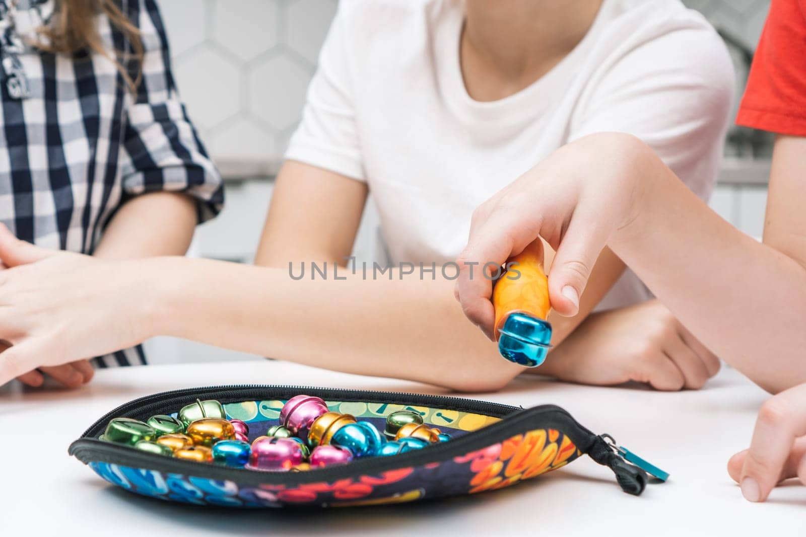 Side view of group of preteen children sitting at white table, catching colorful round metal bells with orange plastic tweezers on soft bright textile tray. Board game, entertainment, development.