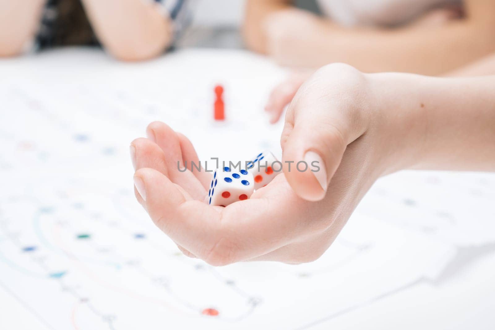 Cropped photo of group of children sitting at table, playing tabletop game, holding white dices over white board with numbers, lines, thinking, counting moving. Board games. Soft focus. Close-up.