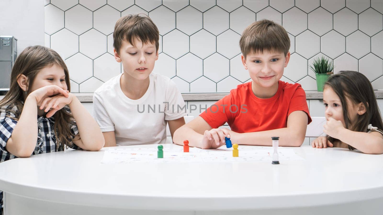 Portrait of group of excited children friends sitting at white table in kitchen at home, playing tabletop game, moving colorful figures on white board with numbers, tracks. Board games, entertainment.