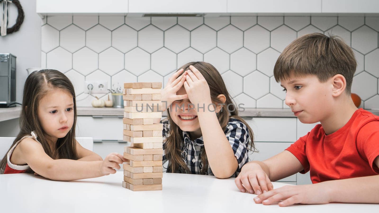 Group of focused children playing board game Jenga on table in kitchen at home building tower made of wooden blocks beams. Worried girl cover eyes with hands waiting friend pull detail. Entertainment.