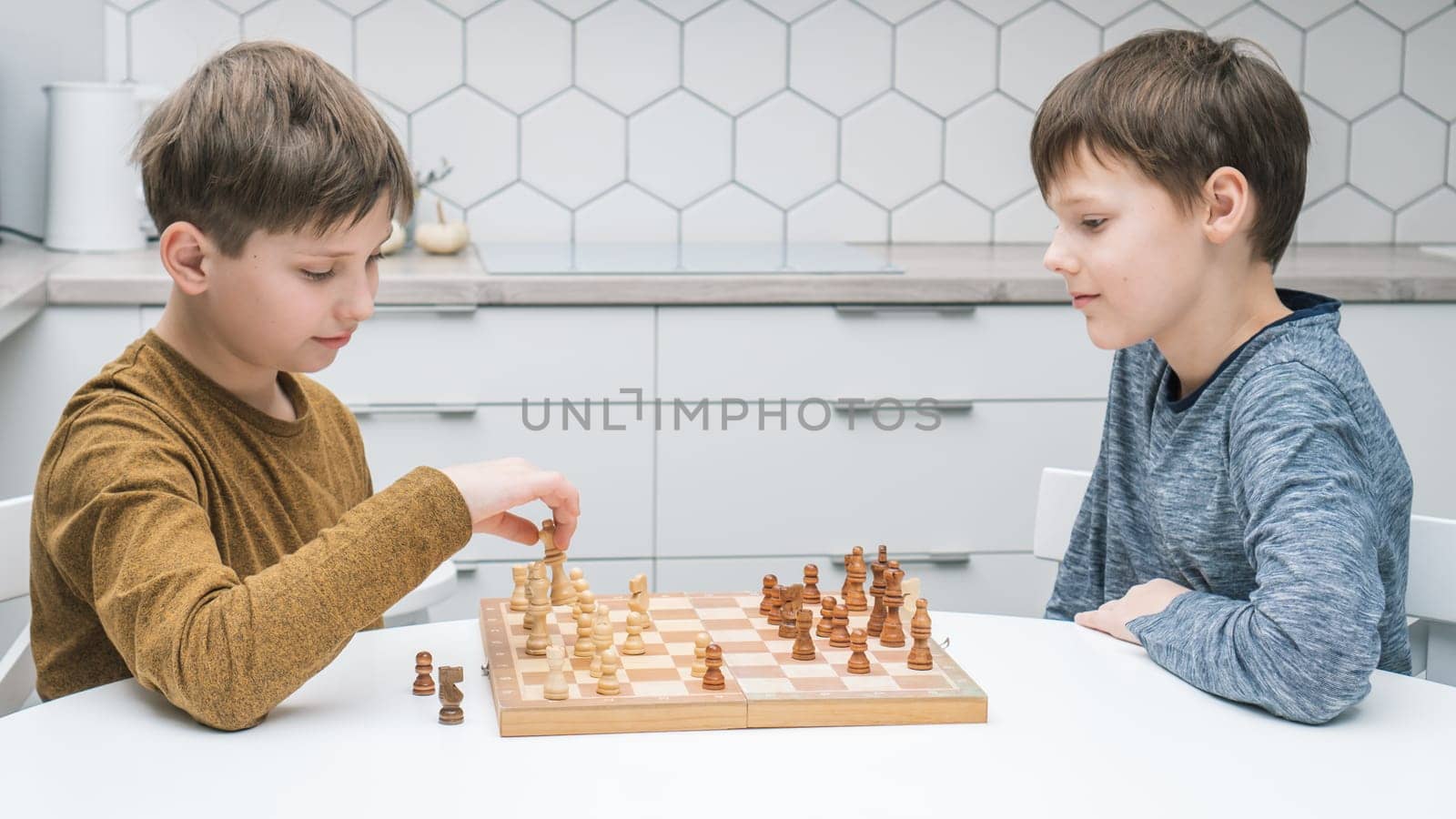 Side view of two calm focused preteen boys friends brothers sitting at white table in kitchen at home, playing chess on wooden board, competing. Board games, development, education, leisure, hobby.