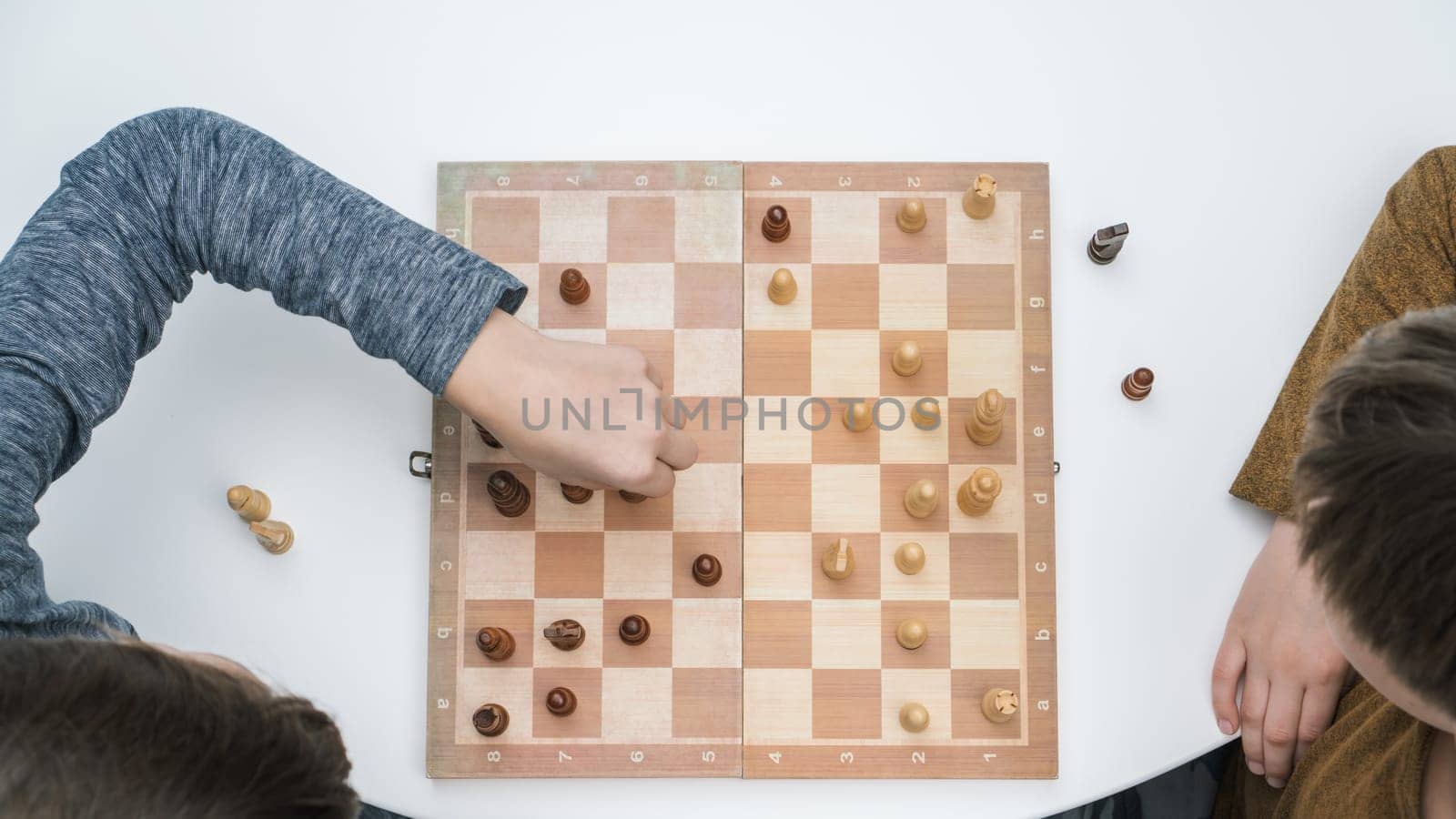 Top view of two focused preteen boys friends brothers sitting at white table, playing chess on wooden board, thinking, building strategy, holding figure with hand. Board games, development, education.