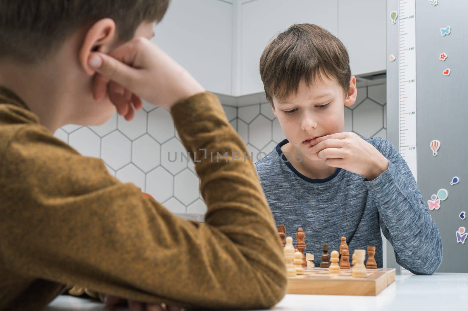 Portrait of two concentrated preteen boys friends brothers sitting at table in bright kitchen at home, playing chess on wooden board, thinking, building strategy. Board games, development, education.