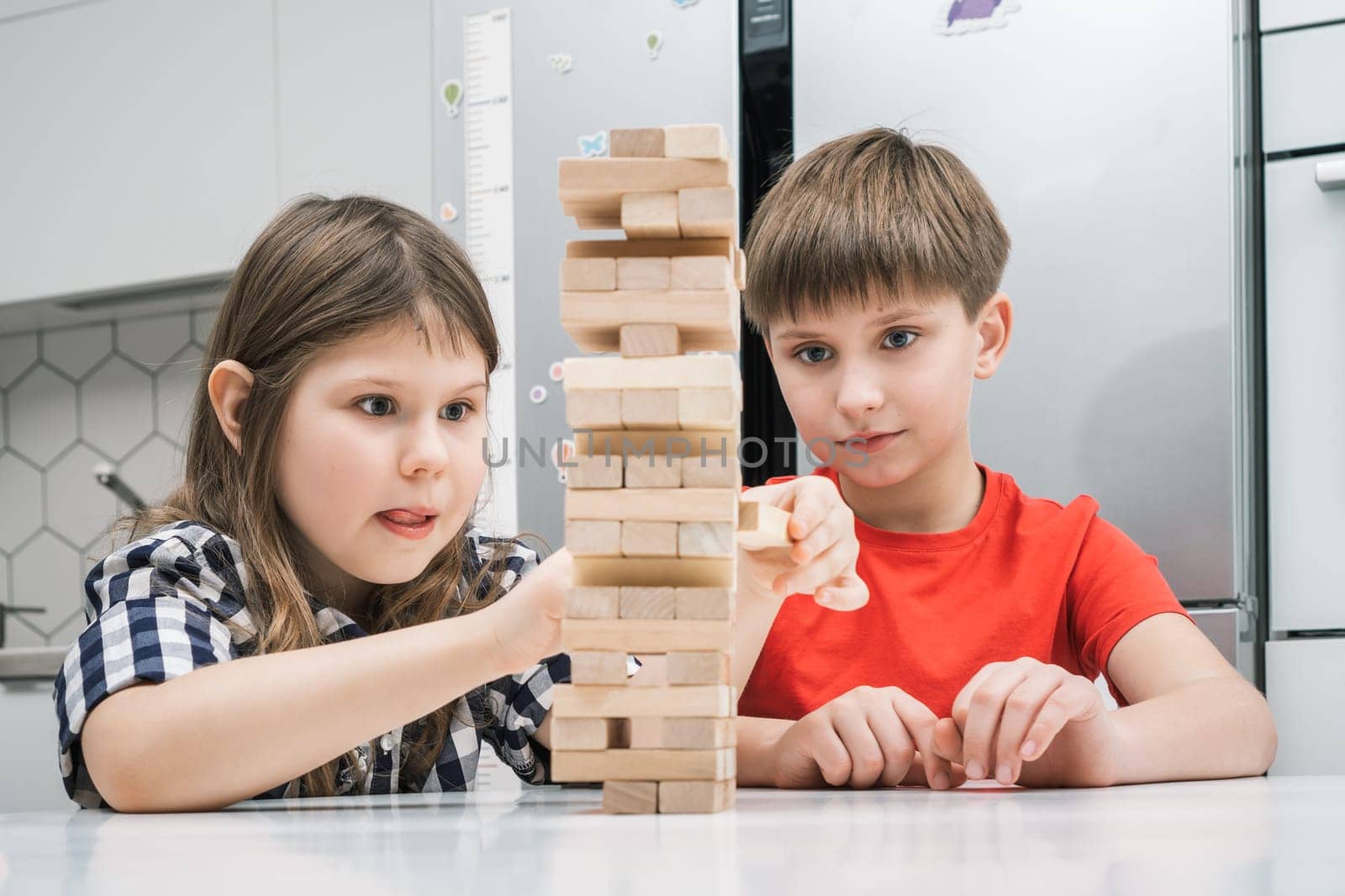 Two children playing board game Jenga on table in kitchen at home, building tower made of wooden blocks. Focused girl with tongue out pulling detail. Board games, hobby, entertainment, education.