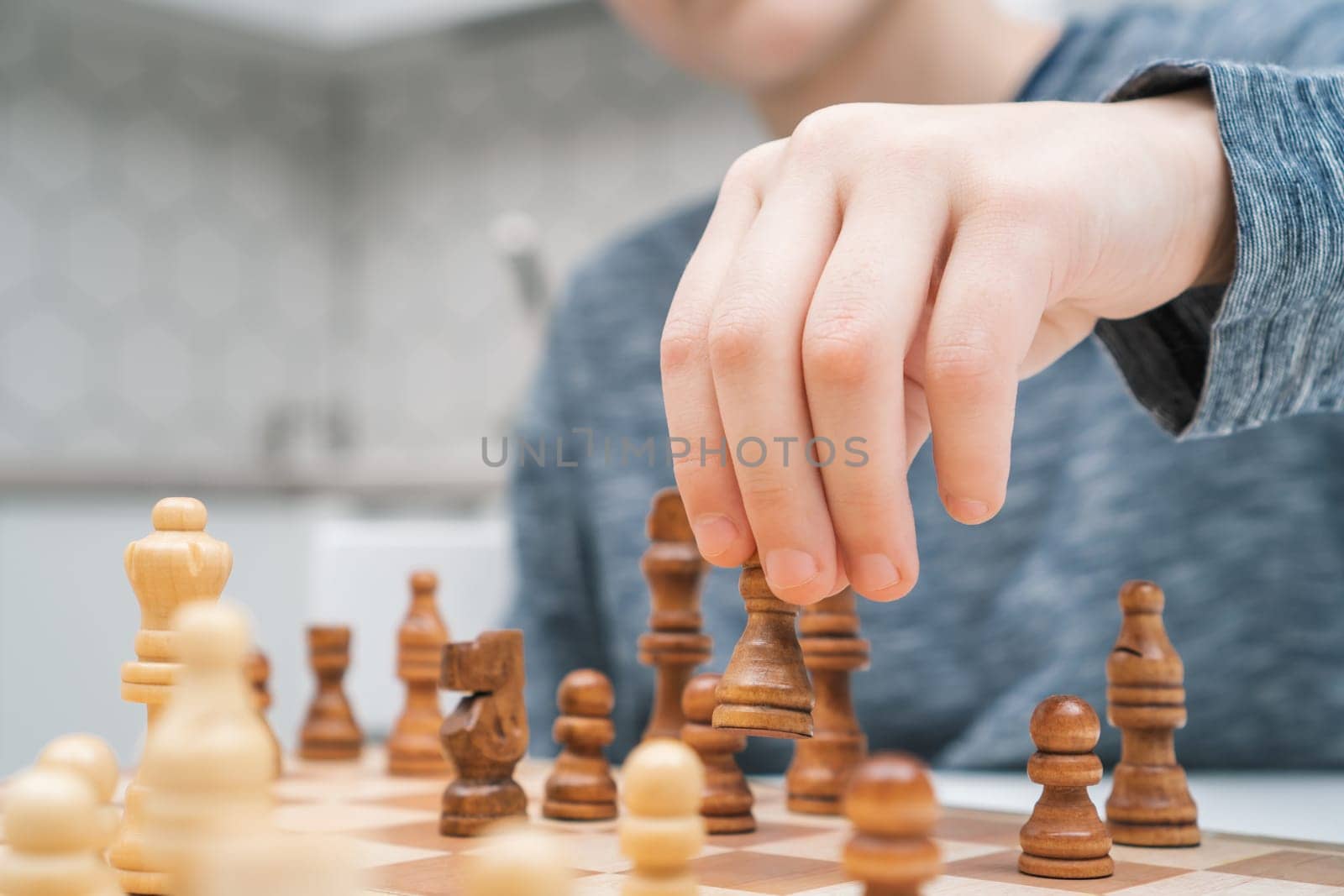 Cropped photo of preteen schoolboy sitting at table in kitchen at home, playing chess on wooden board, planning, building strategy, holding dark figure with hand. Board games, development, education.