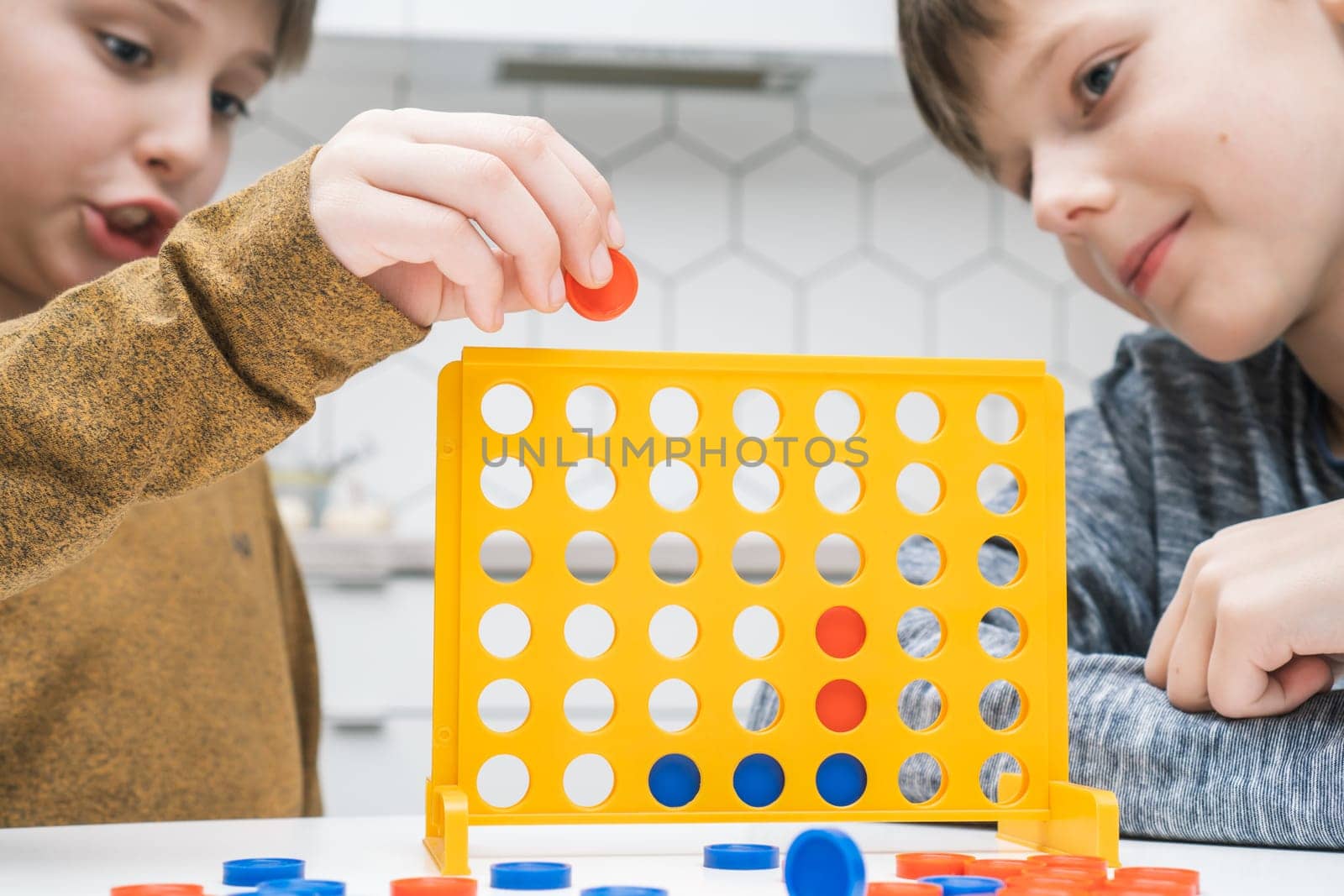 Portrait of two preteen boys friends children sitting at white table in kitchen at home, playing board game connect four 4 in line, inserting checkers into yellow plastic grid. Board games, education.