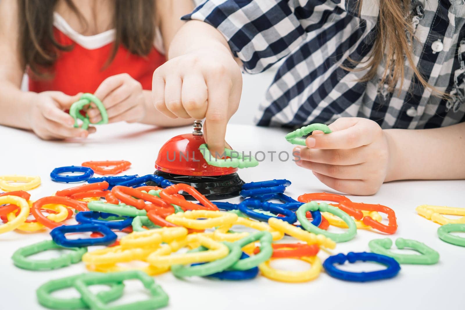 Cropped photo of group of girls friends children standing at white table, playing board game ring near bell, joining buckling holding different colorful geometric shapes figures in chain. Board game.