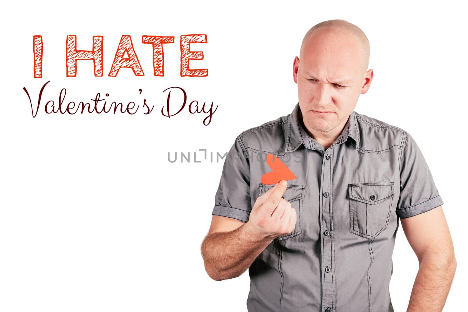 Handsome frowning bald middle-aged man in gray shirt hold red paper heart with two fingers and inscription I hate Valentines day isolated on white background. Valentine's day, love, emotion concept