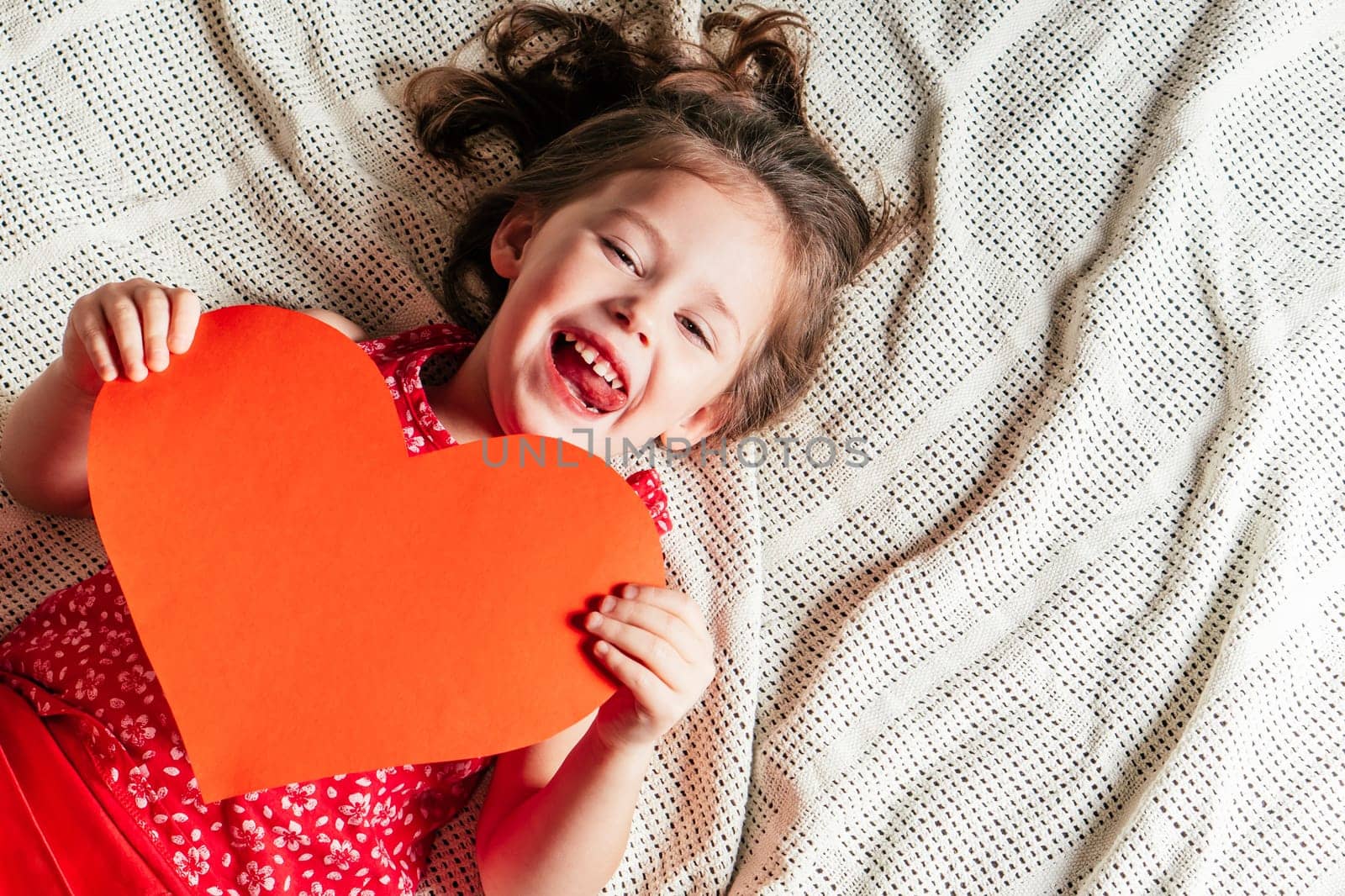 Portrait of smiling preschool cute little girl lying on light bedspread holding large heart Valentine card in her hands