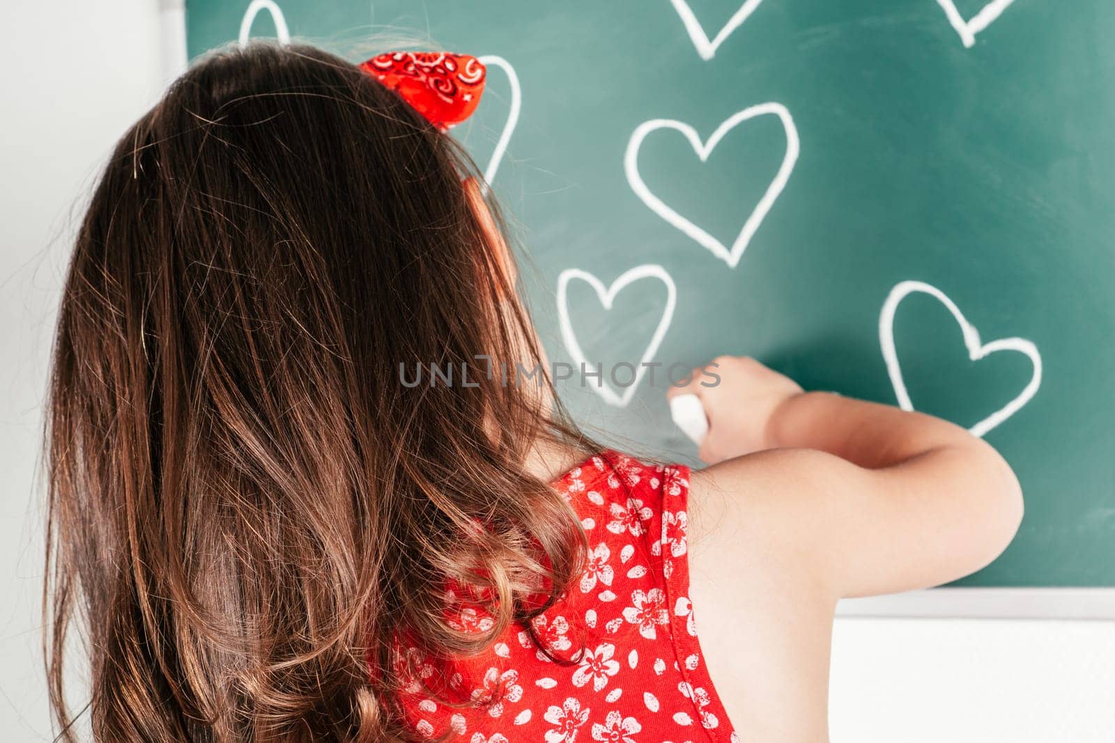 Little girl with long curly hair in red clothes standing back, drawing hearts with chalk on green blackboard on white background. Family, love, holidays, birthday, Mother's day concept. Copy space.