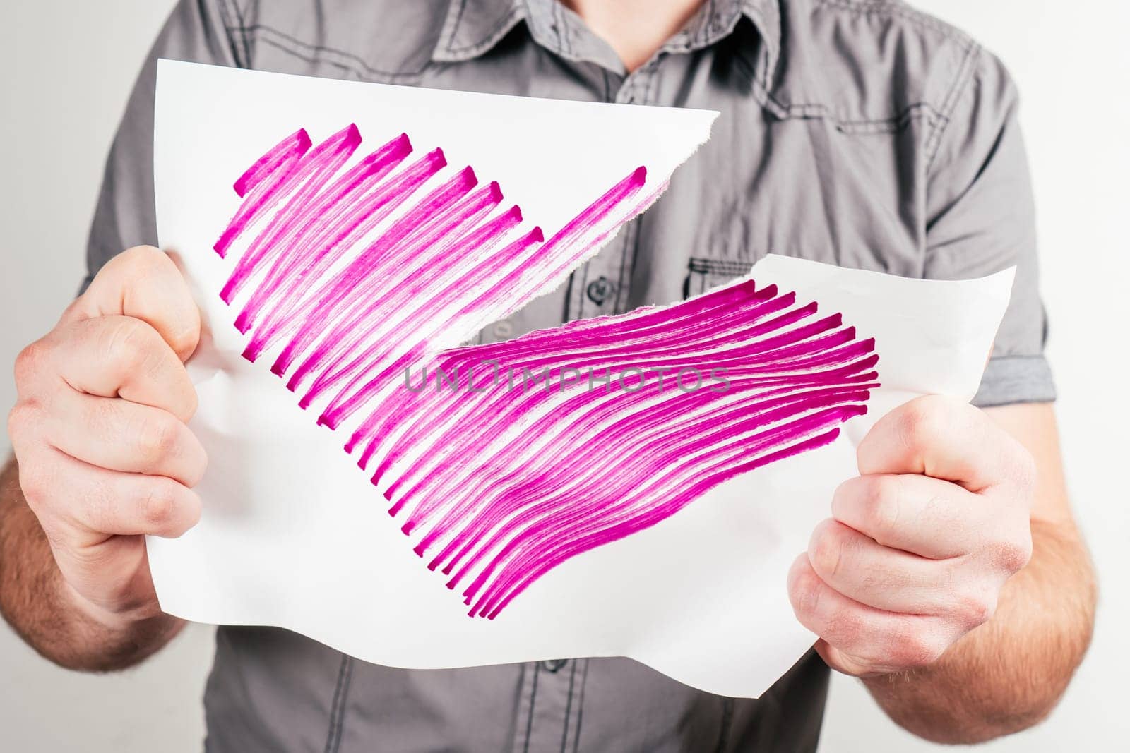 Unrecognizable man in gray shirt tear pink heart drawn on sheet of paper in different directions on white background. Valentine's day, hatred, dislike, broken love, toxic relationships, emotion